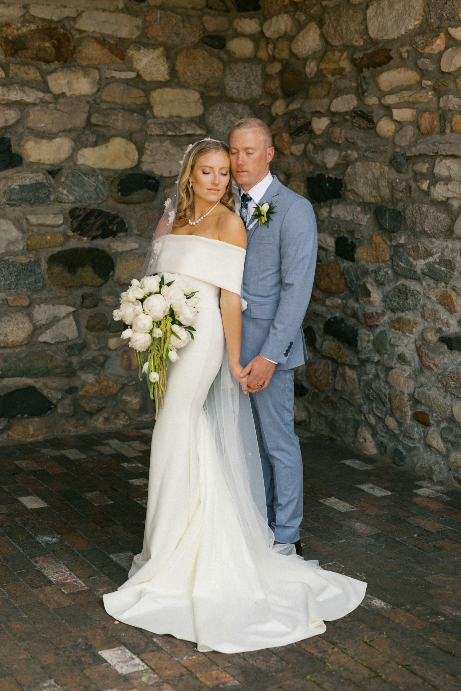 A wedding couple poses against a rustic stone wall backdrop with a stunning white bouquet.