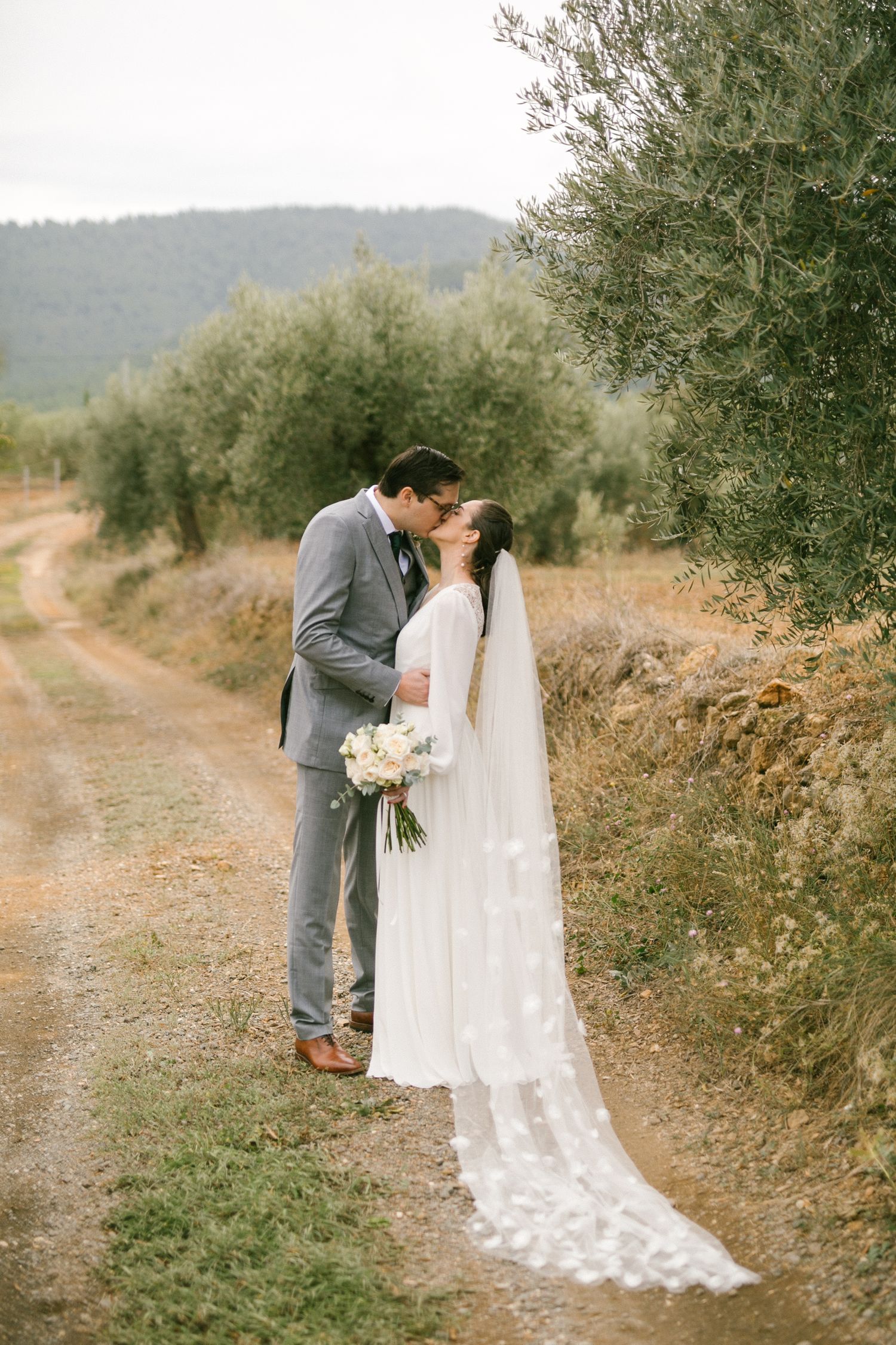 A bride and groom share an intimate moment on a dirt path surrounded by olive trees in a rustic outdoor setting in Madrid, Spain.