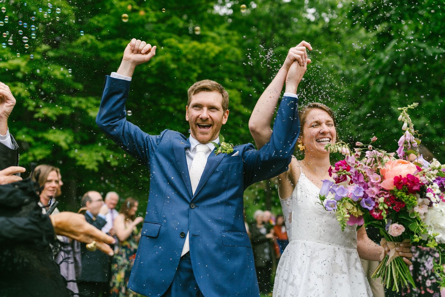 Joyful newlyweds celebrate with raised hands during outdoor garden wedding ceremony while holding vibrant pink bouquet.