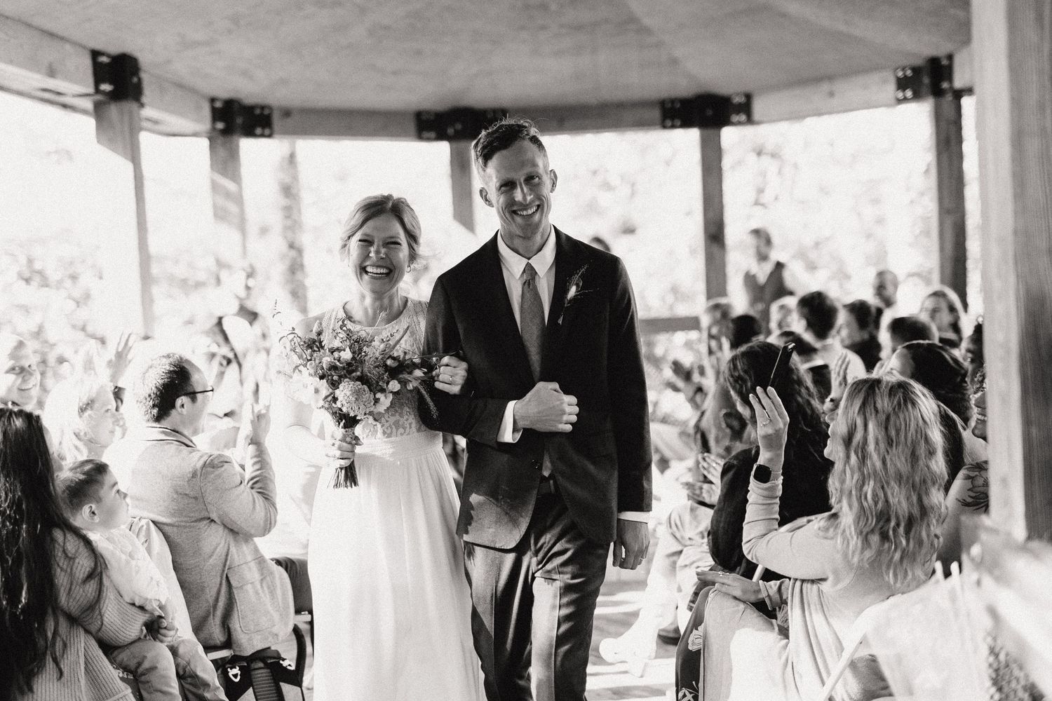 Black and white capture of joyful wedding ceremony exit in rustic wooden pavilion.