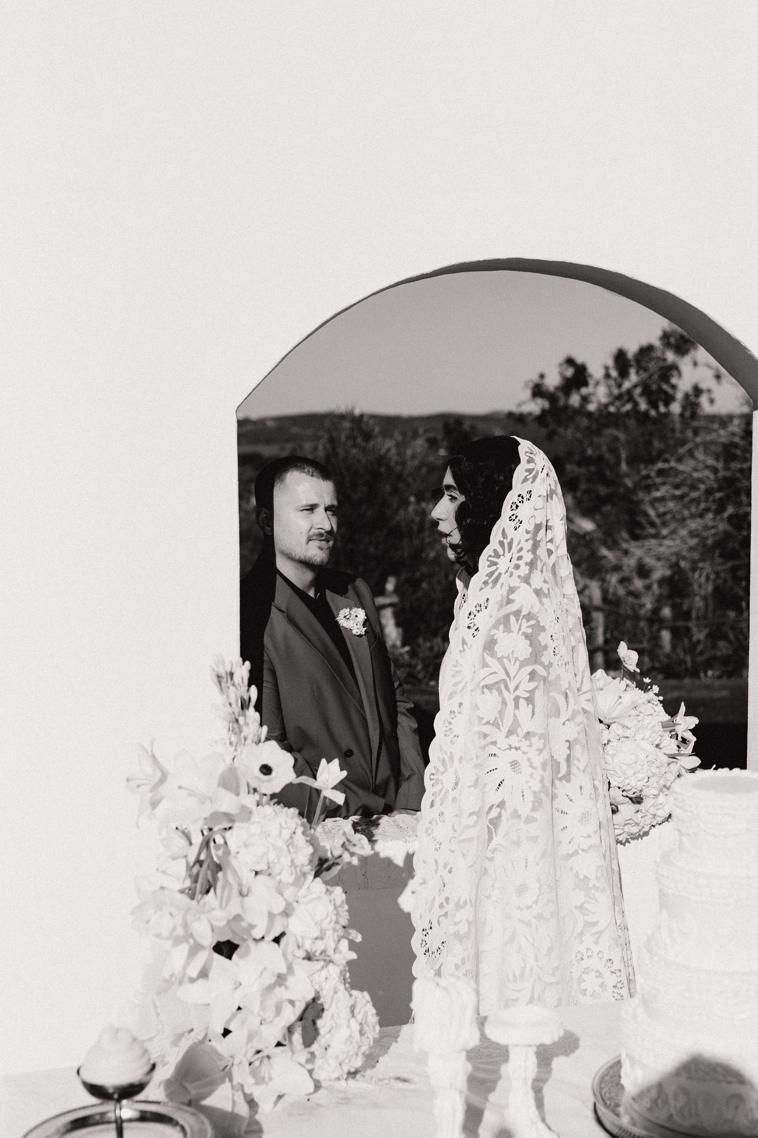 Black and white wedding photo of bride in lace veil and groom exchanging vows at elegant outdoor ceremony.