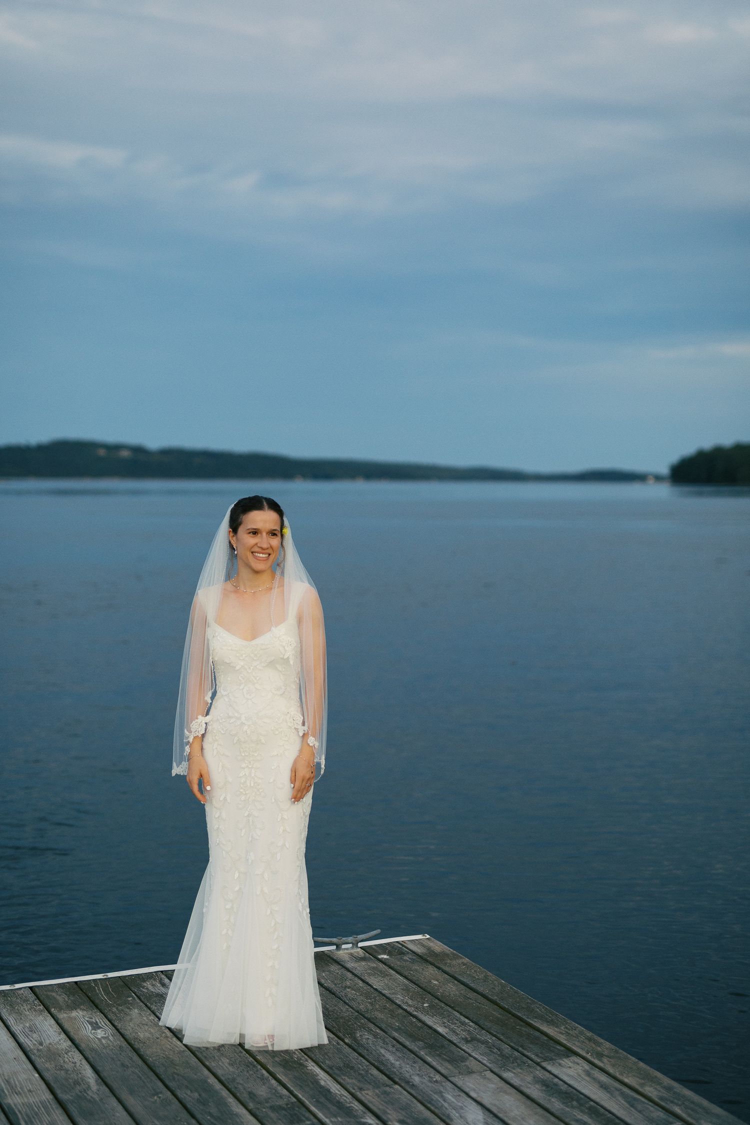 Bride in white lace gown and veil stands on wooden dock overlooking the calm Kennebec River at dusk.