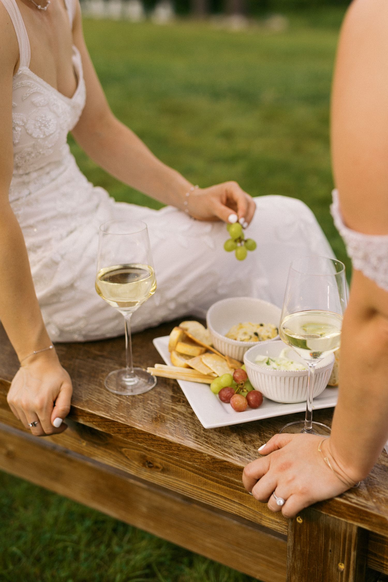 Two women in white dresses enjoy a picnic with white wine, grapes, cheese, and strawberries on a wooden bench outdoors.