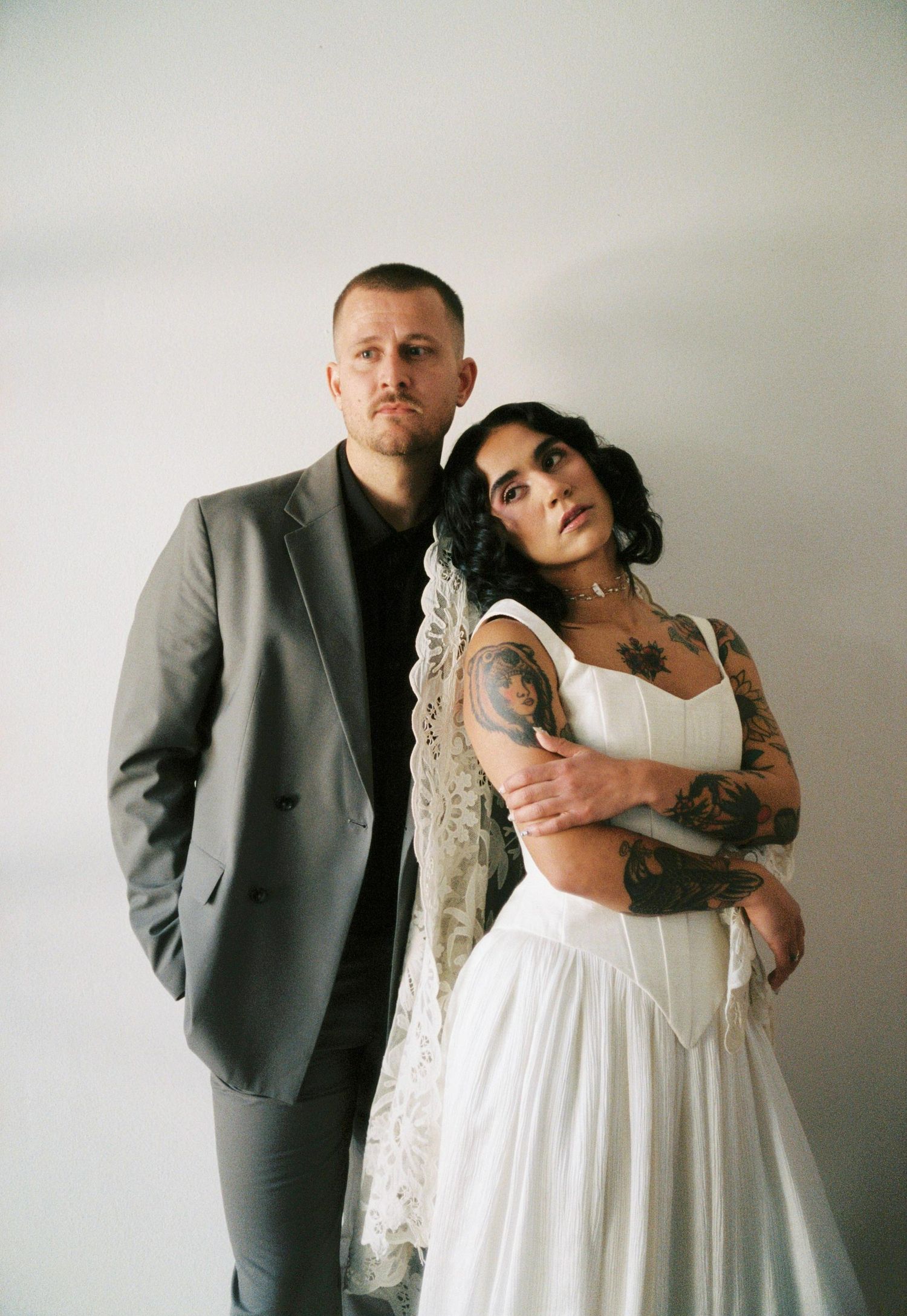 Tattooed bride in white dress leaning on groom in grey suit against a white wall.