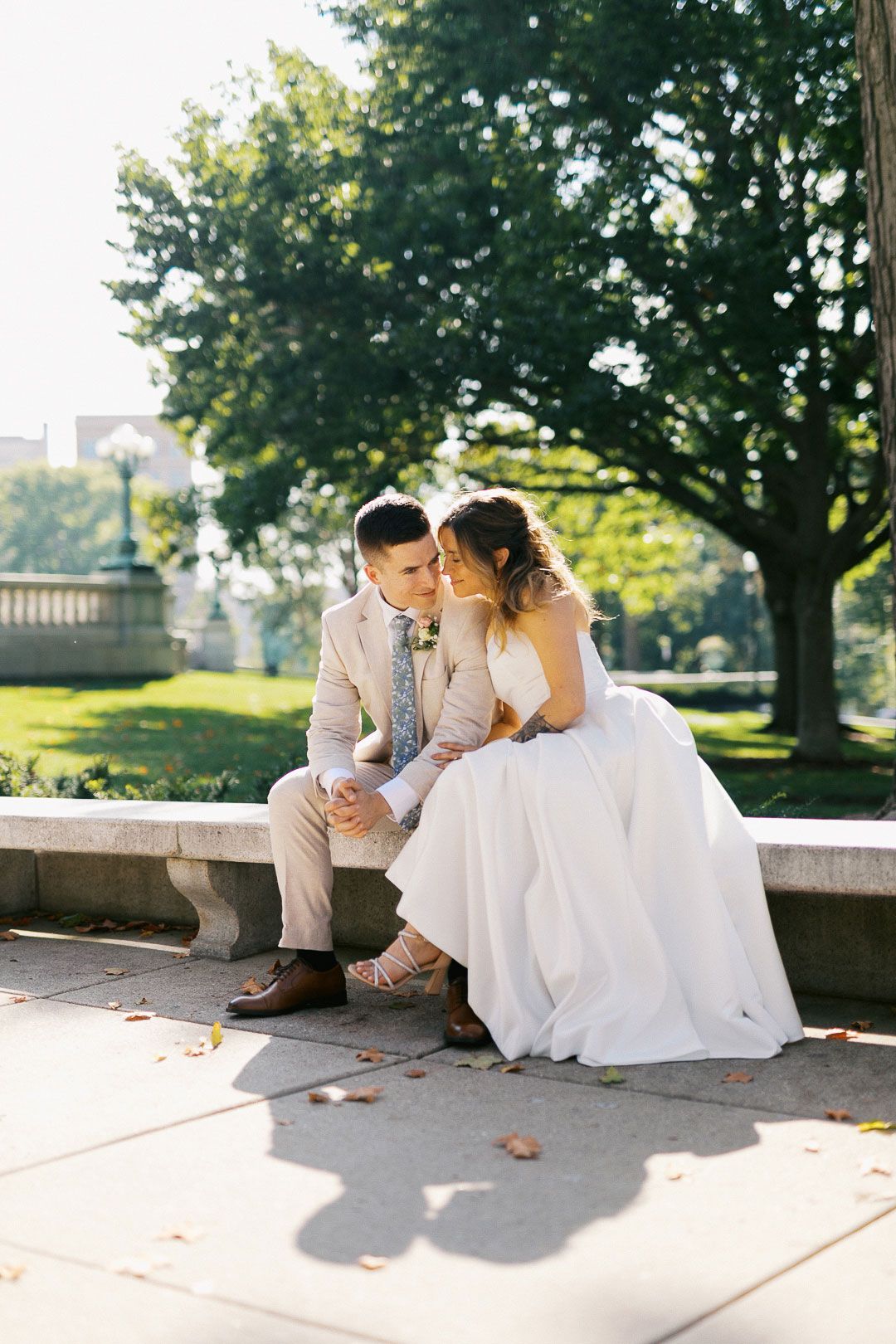 Bride and groom sit closely together on stone bench in sunny park setting.