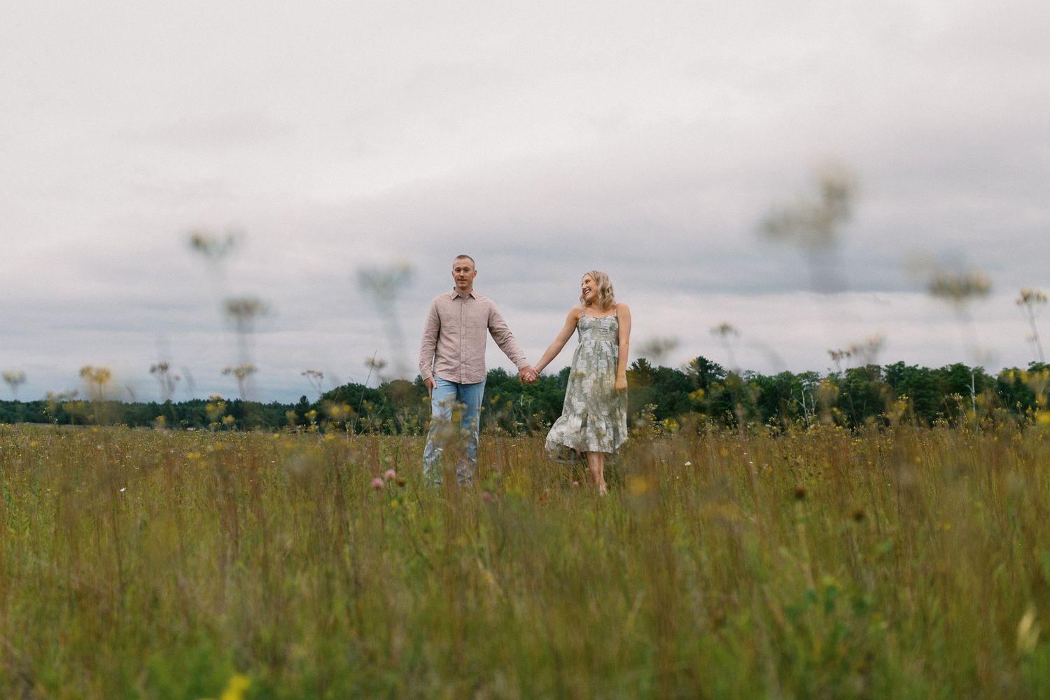 Couple holding hands while walking through a lush green field under a cloudy sky.