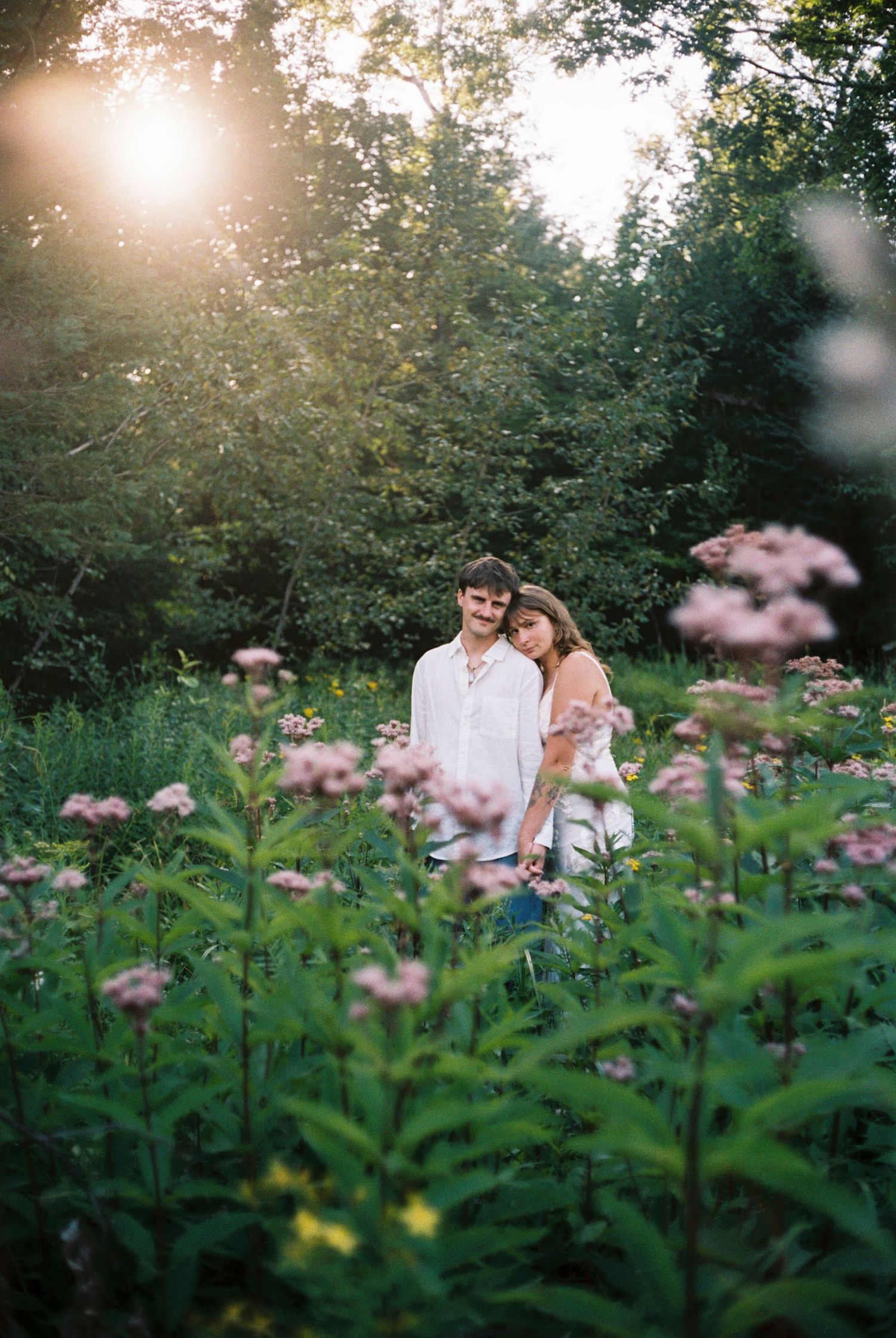 Couple embracing in a sunlit wildflower meadow surrounded by lush green trees during golden hour.