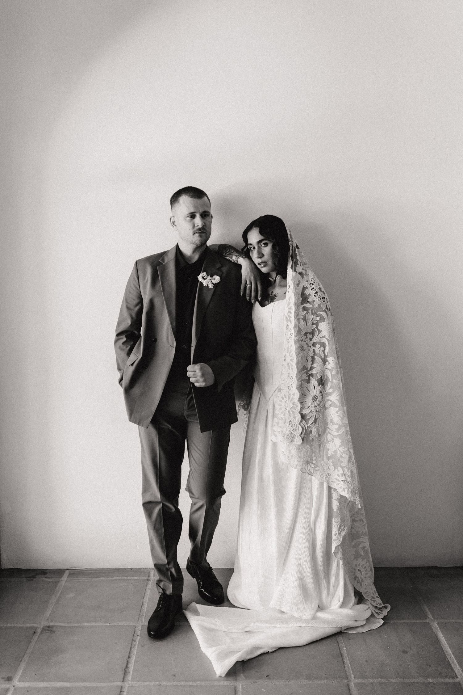 Black and white portrait of bride and groom posing together, bride wearing lace veil wedding dress, groom in grey suit.