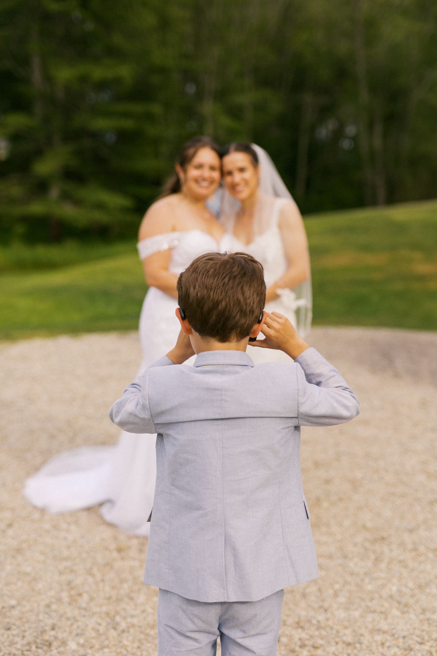 A young boy in a gray suit photographs two brides in white wedding dresses posing on a gravel path outdoors.