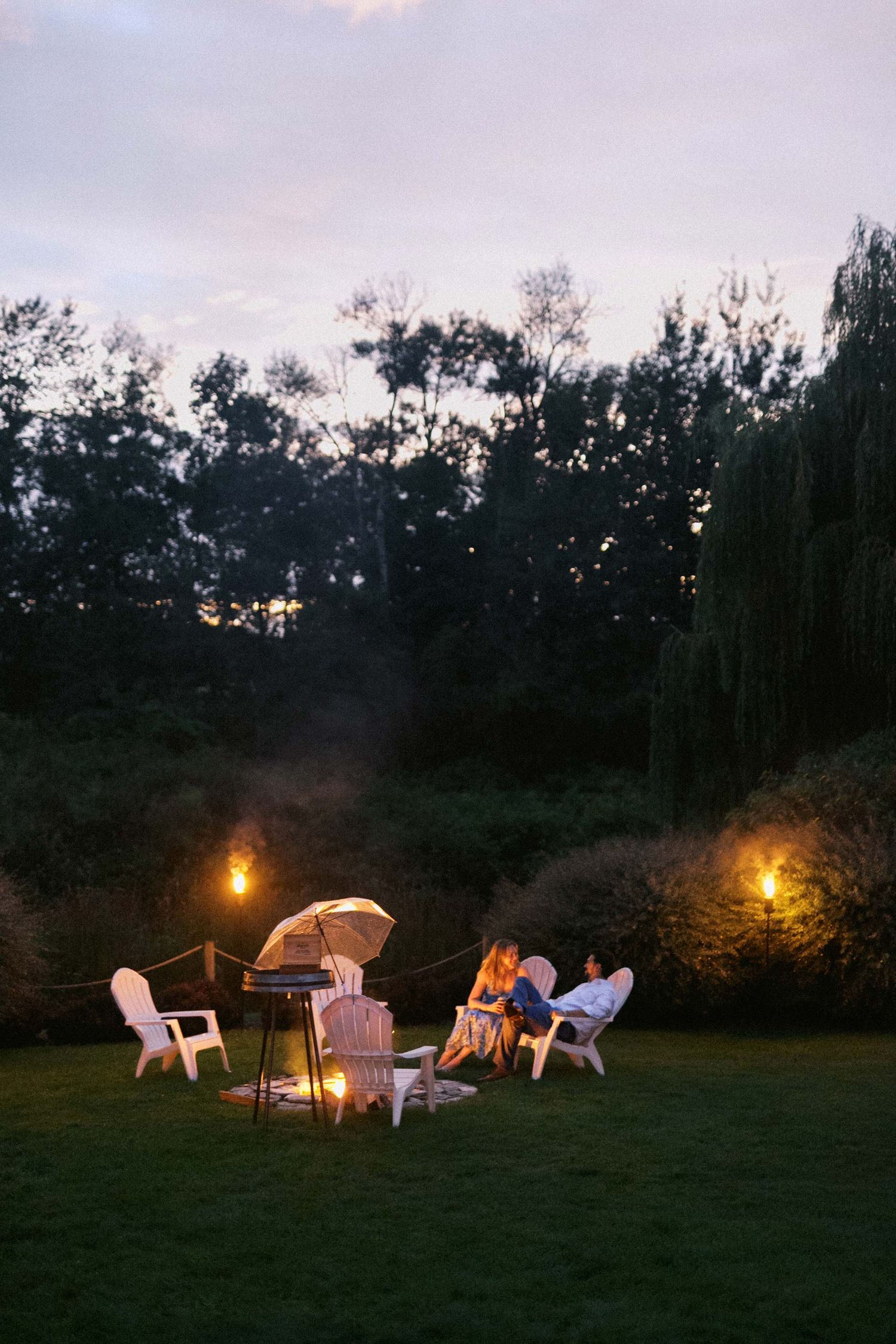White wicker chairs arranged around a glowing fire pit at dusk create a cozy outdoor seating area.