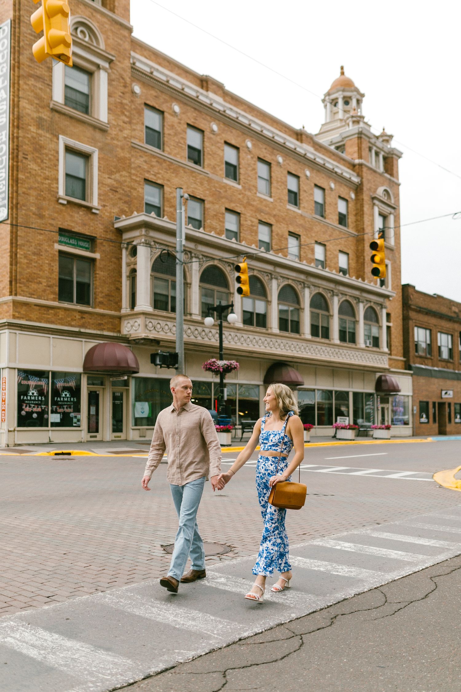 Two people cross a brick-lined street with historic architecture and storefronts visible behind them.
