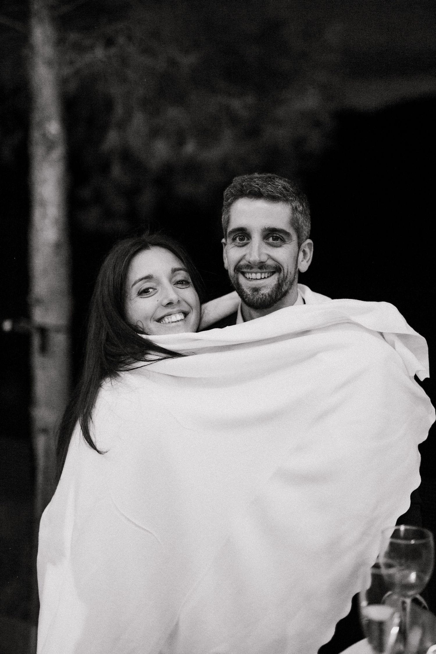A couple wrapped in a white blanket share a tender moment together in black and white photography.