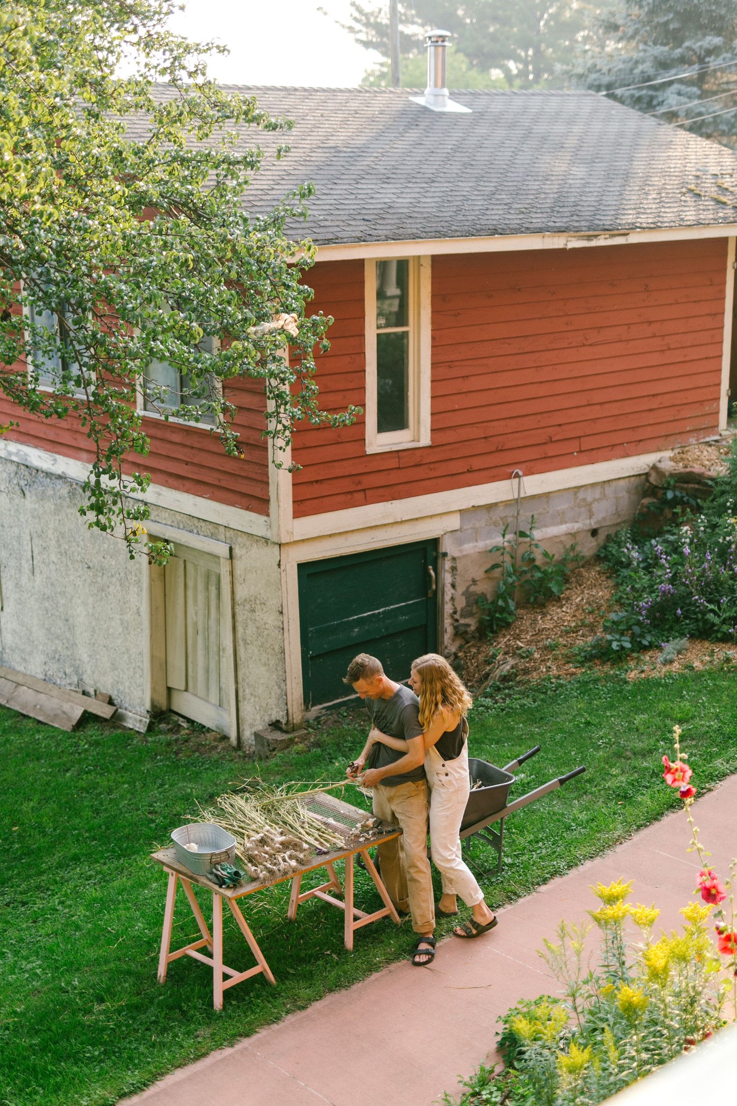Couple grilling outdoors beside a red house with garden flowers in bloom.