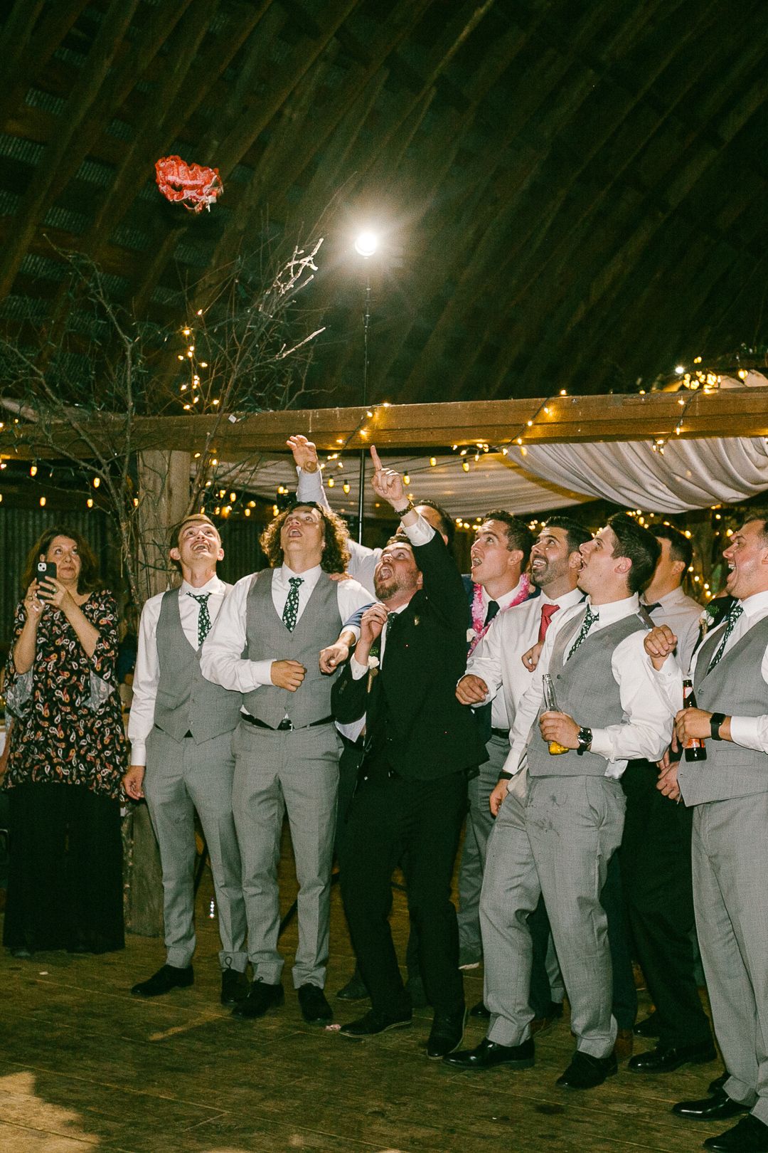 Wedding reception party guests in grey suits watching a bouquet toss under string lights in a dimly lit venue.