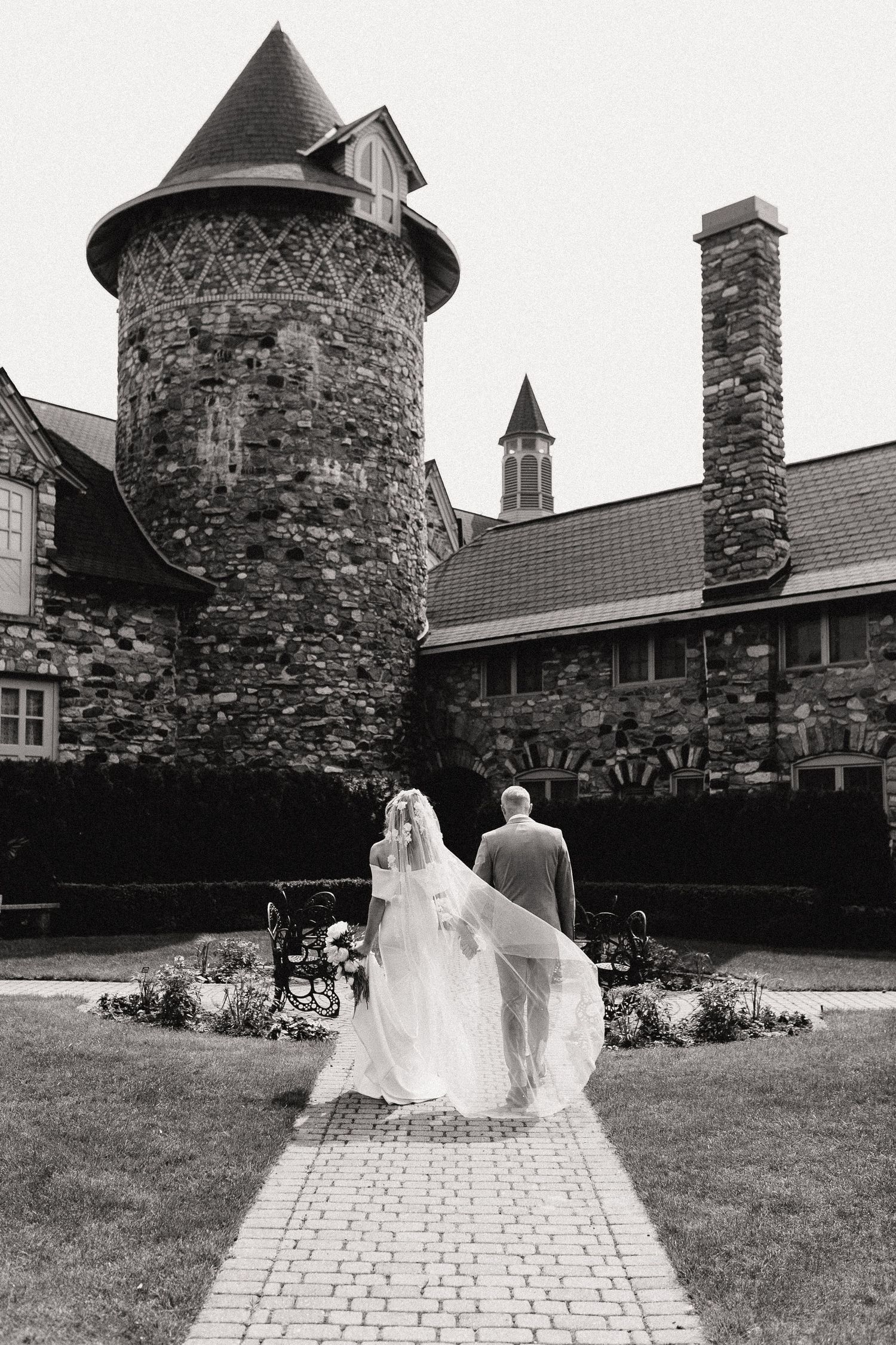 A couple walking away on brick path towards a stone castle-like building in black and white.