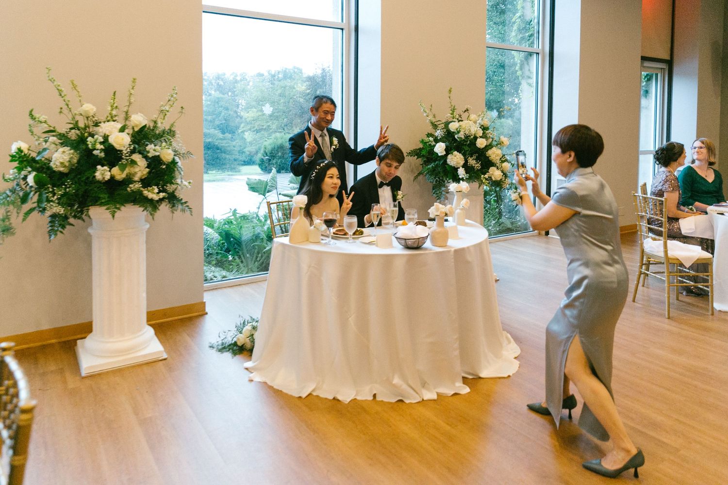 Elegant wedding reception with floral arrangements, guests toasting at a sweetheart table in a bright venue.