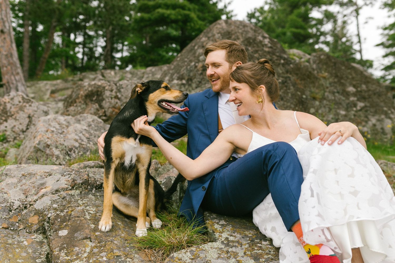 A couple in wedding attire sits on a rocky outcrop petting their black and tan dog in a scenic outdoor setting.