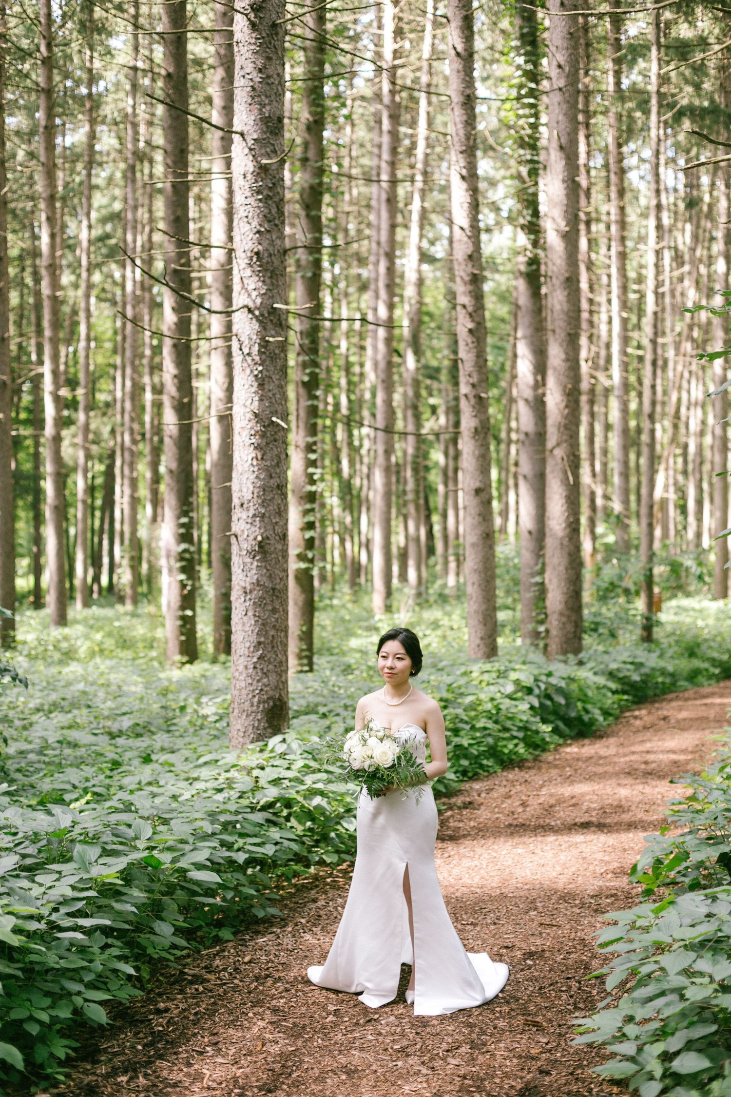 Elegant bride in a white strapless gown holding a bouquet on a forest path surrounded by tall trees.
