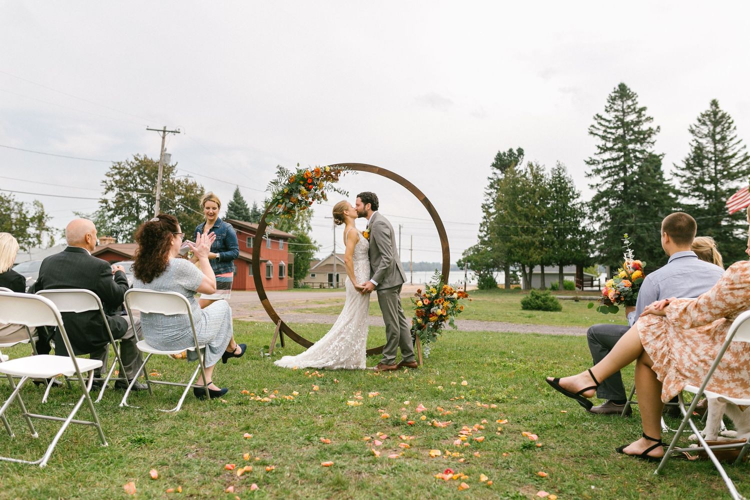 An outdoor wedding ceremony with a wooden arch decorated with flowers takes place on a grassy field with pine trees in the background.