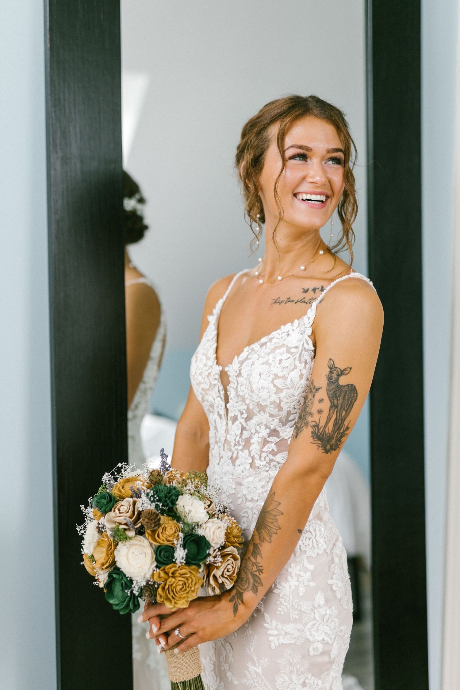 A bride in a lace wedding dress smiles while holding a peach and cream bouquet with floral tattoo on her arm.