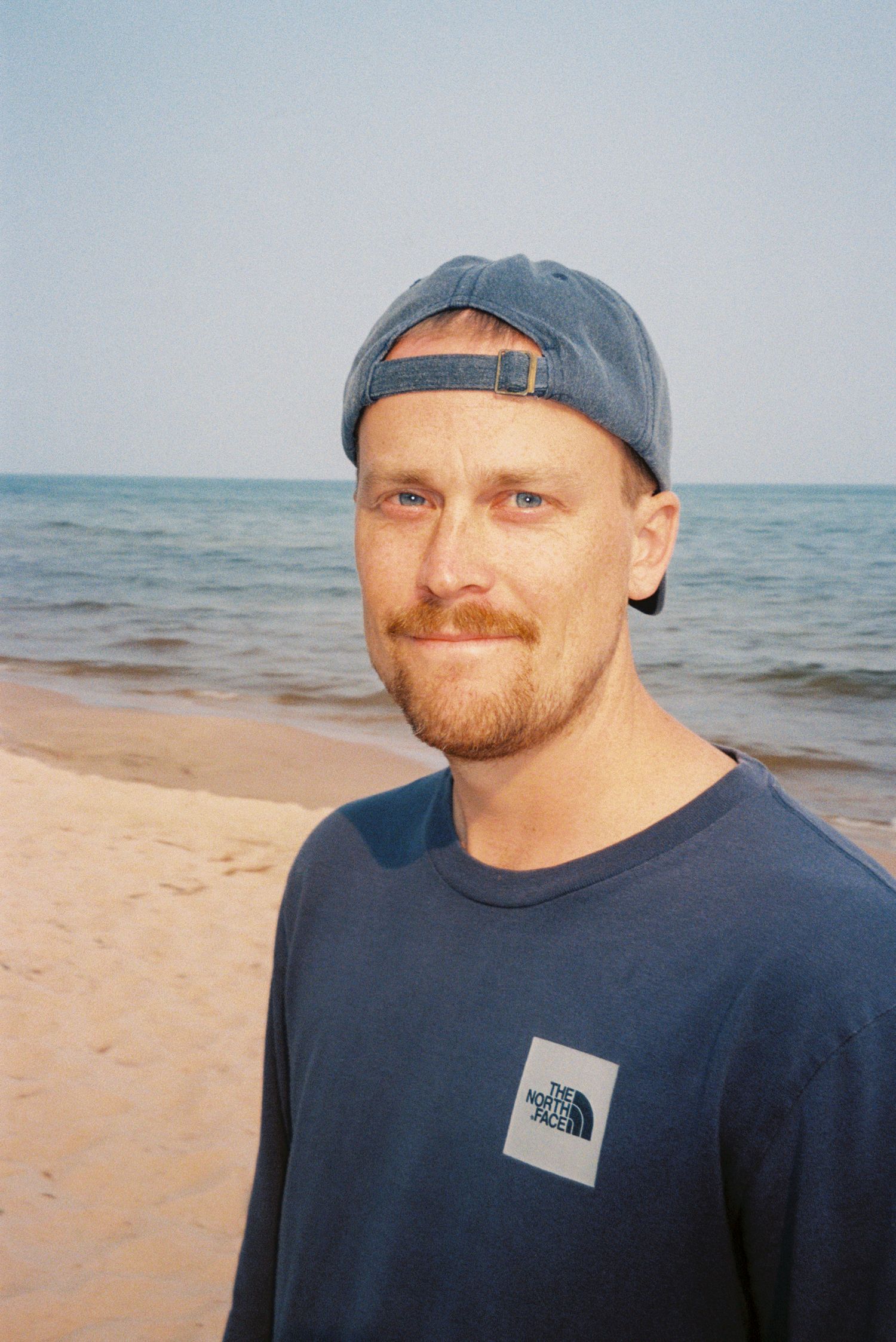 Smiling man in North Face shirt and backward cap poses on sandy beach with ocean behind him.