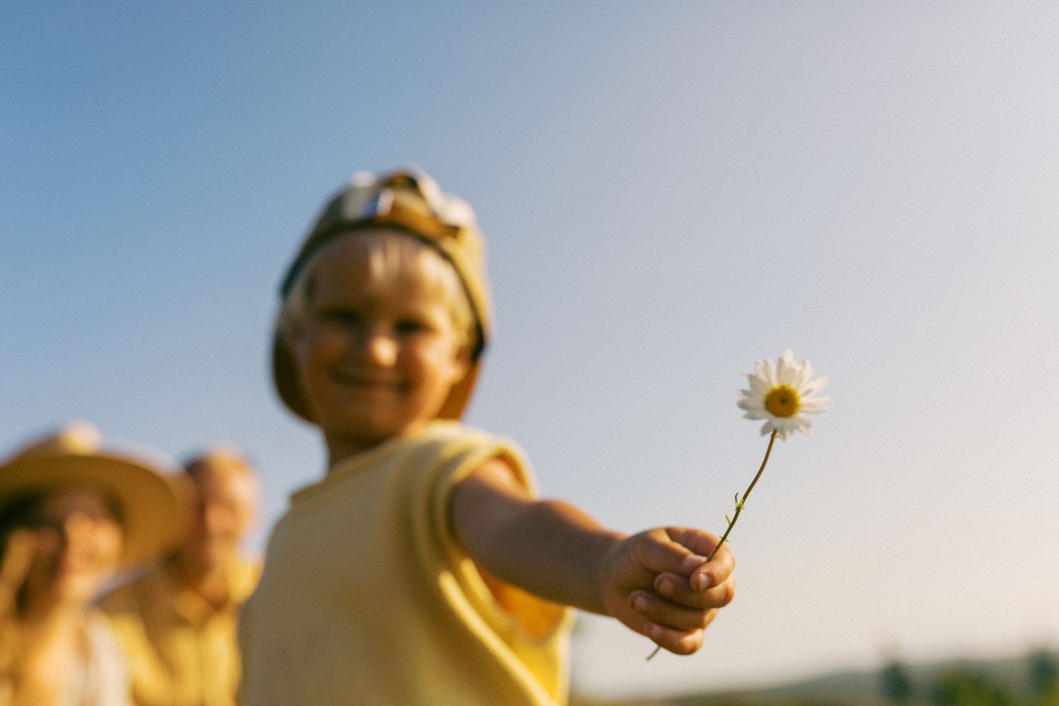 Happy child in yellow outfit extending a daisy flower toward the camera in an outdoor setting.