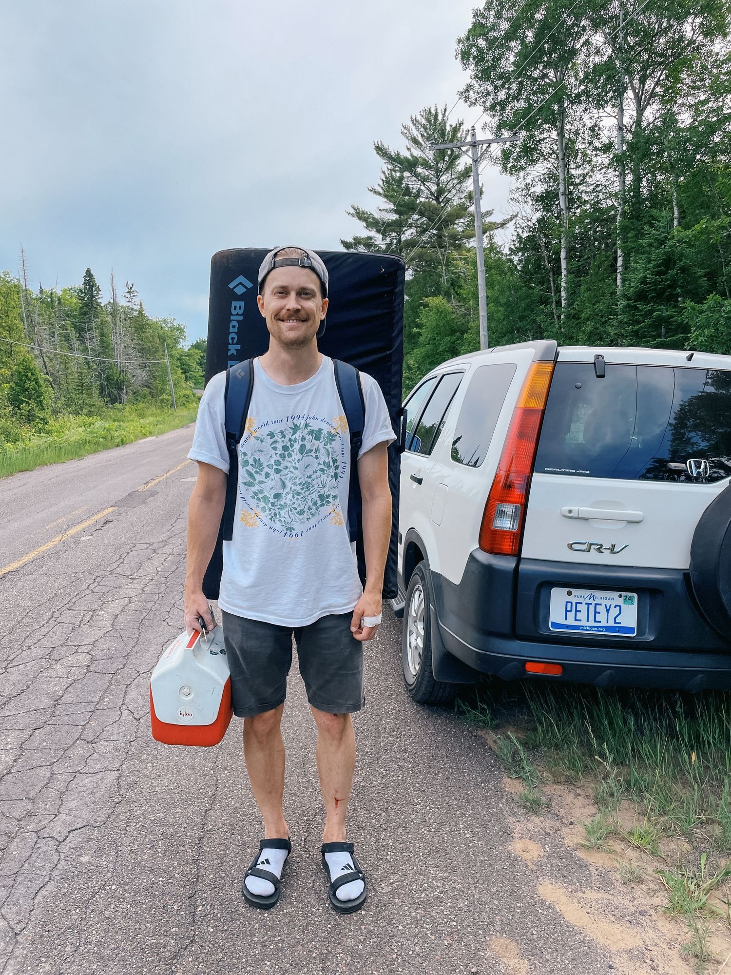 Man with backpack and cooler standing on forest road next to Honda CR-V SUV.