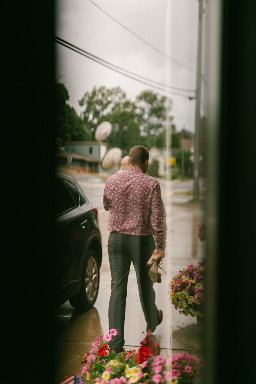 A person bends down to pick flowers on a rainy day, viewed through a window frame.