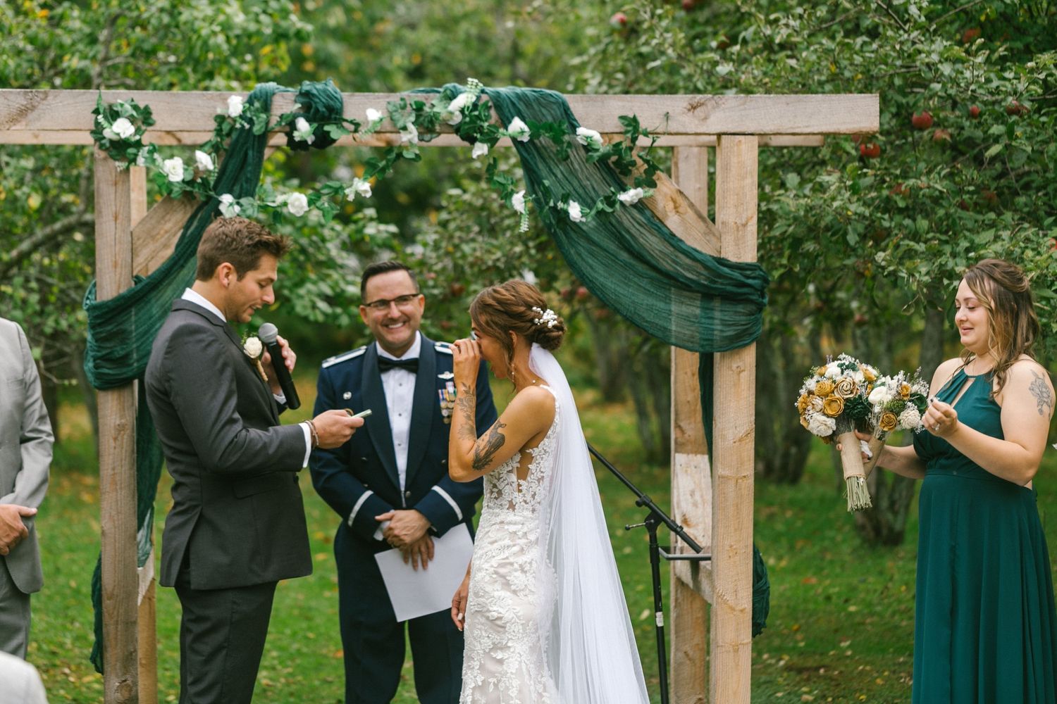 A wedding ceremony takes place under a rustic wooden arch decorated with green fabric and white flowers in a garden setting.