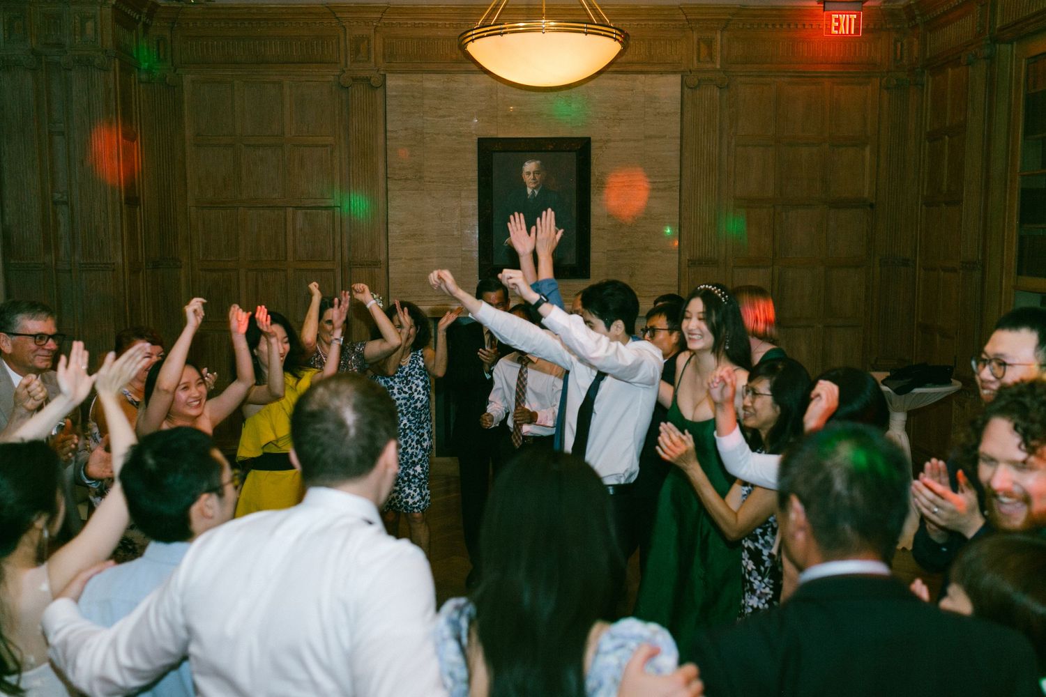 Wedding guests dancing and celebrating at a lively indoor reception in an elegant wood-paneled ballroom.