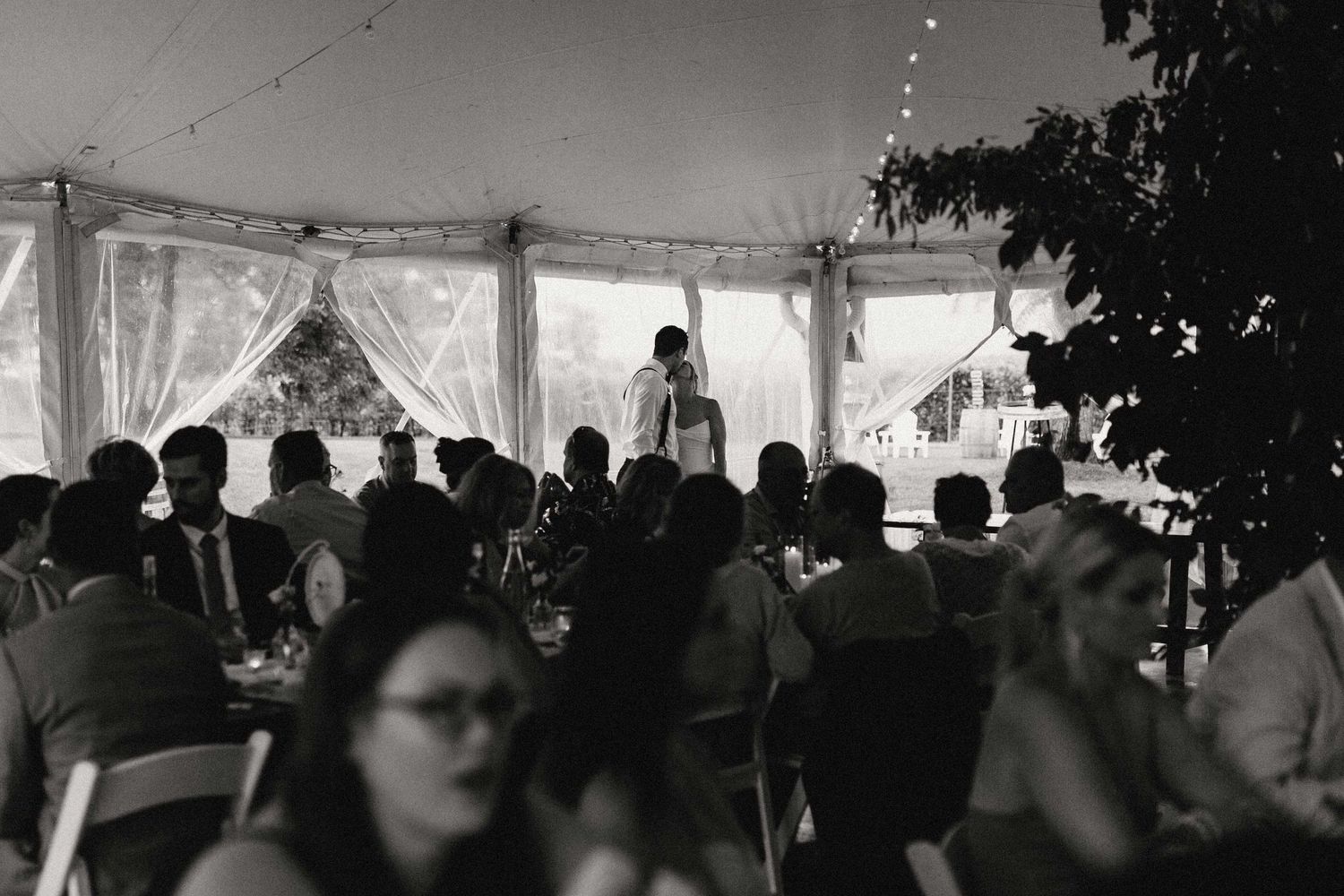 A black and white photograph of guests seated under an elegant reception tent while the bride and groom kiss in the background.