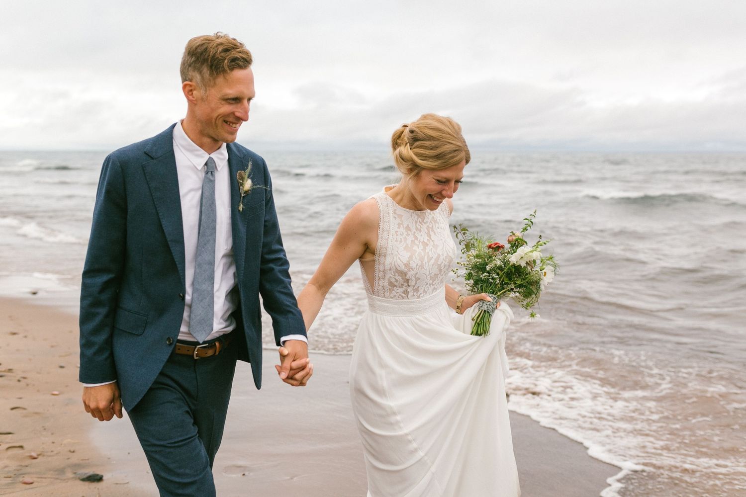 A joyful couple walking on the beach during their wedding, holding hands and carrying a bouquet.