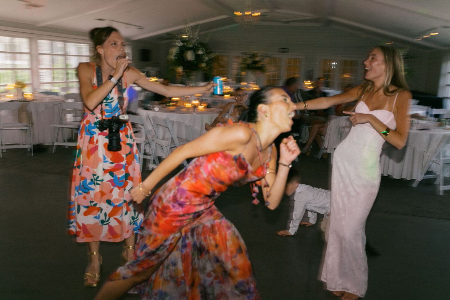 Several people dance energetically on a reception hall dance floor, wearing colorful floral and white dresses.