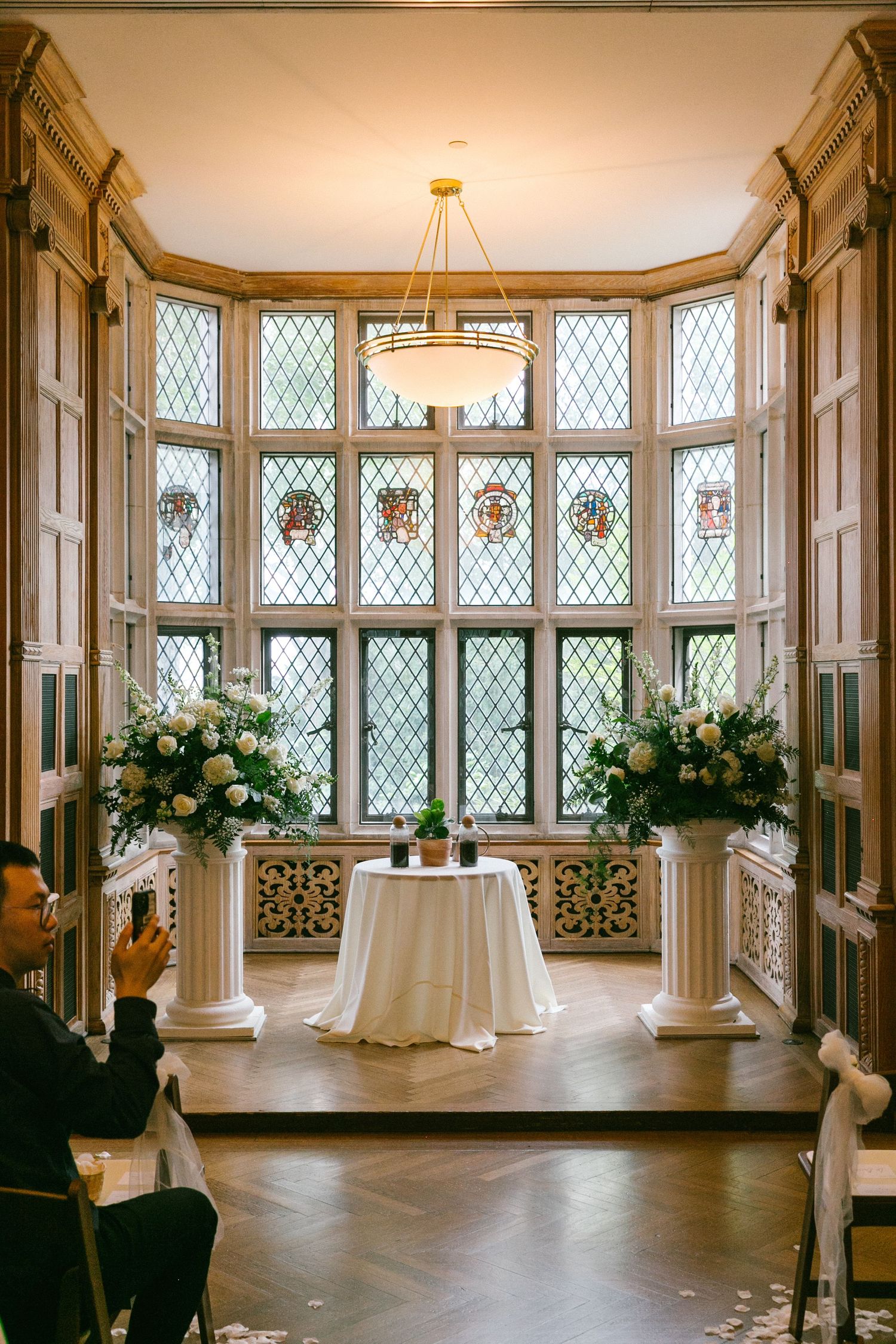 Elegant wedding ceremony setup with white floral arrangements flanking an altar before leaded glass windows.
