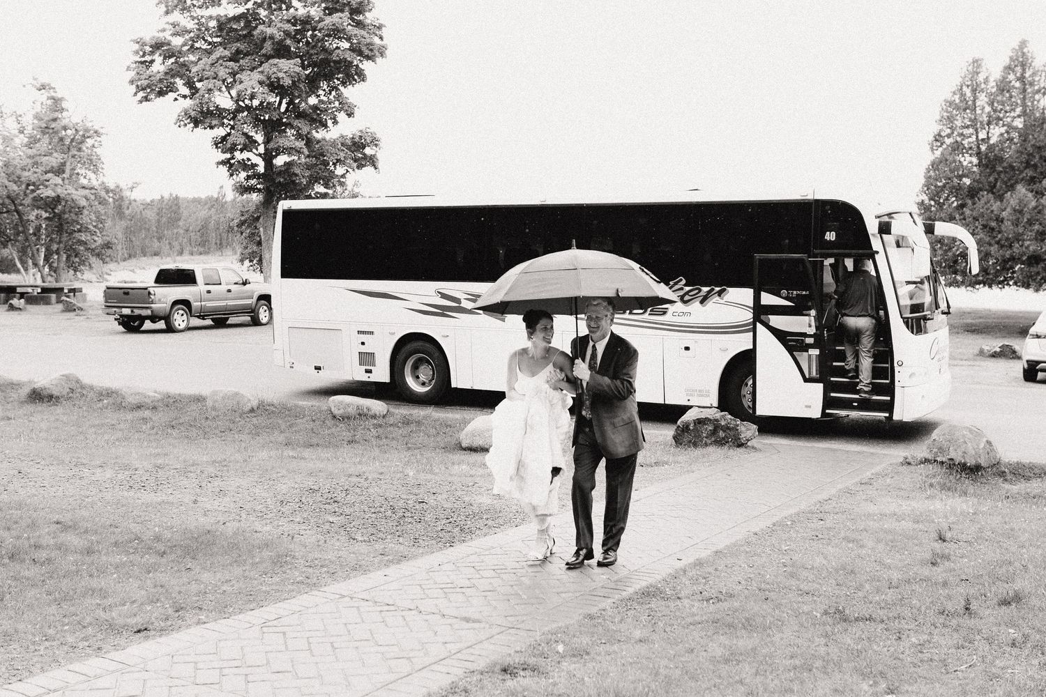 A couple shares an umbrella walking past a parked tour bus in a black and white photograph.