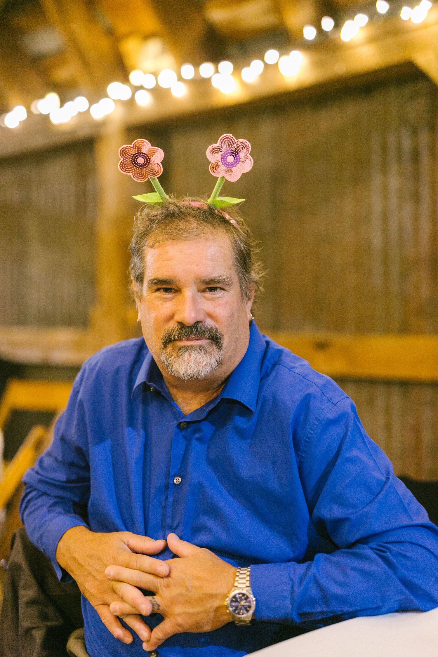 Someone wearing a blue button-down shirt with pink flower decorations in their hair sits in a rustic venue with string lights.