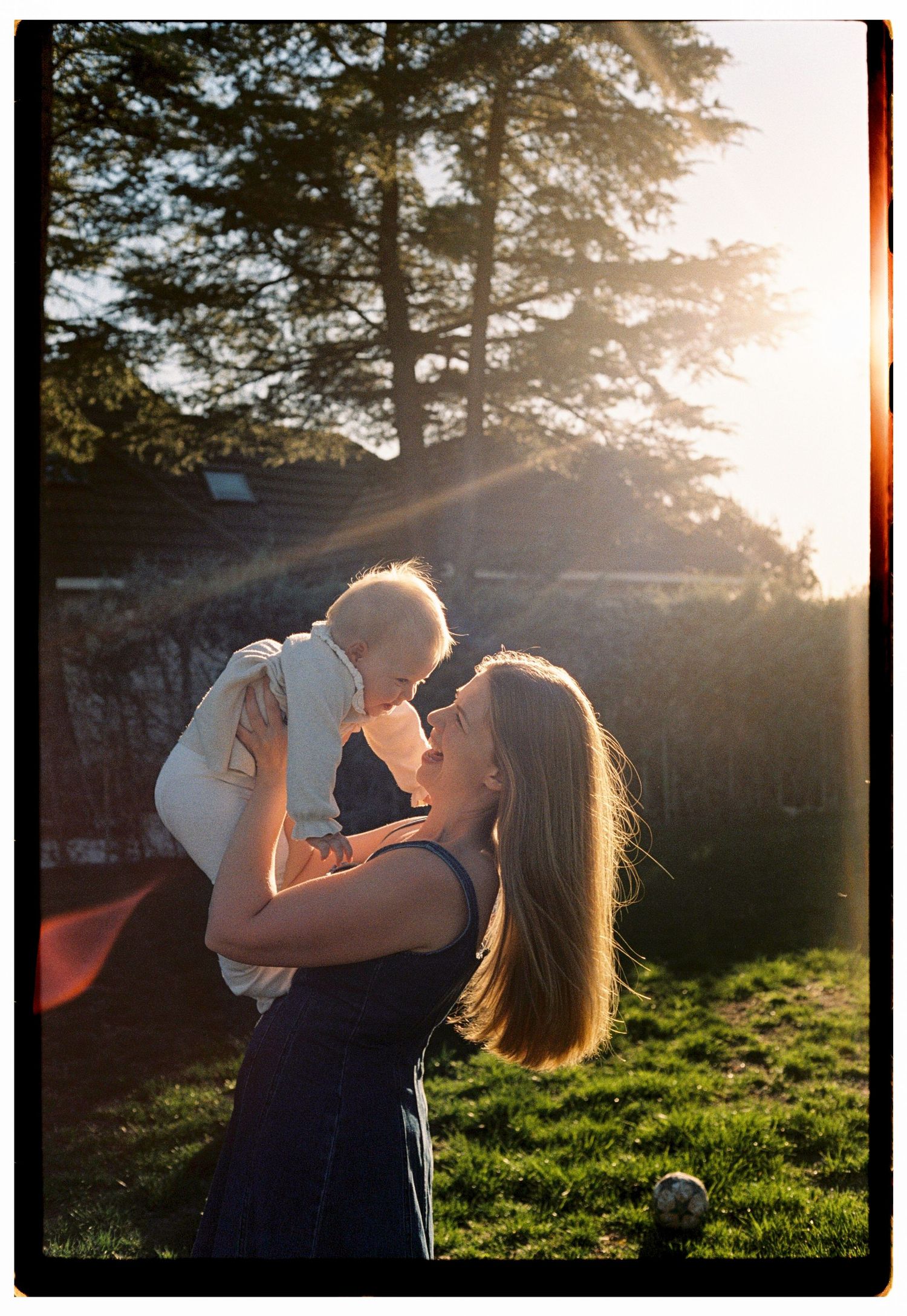 A mother lifts her baby in golden backlit sunlight in a garden, sharing a tender moment outdoors.