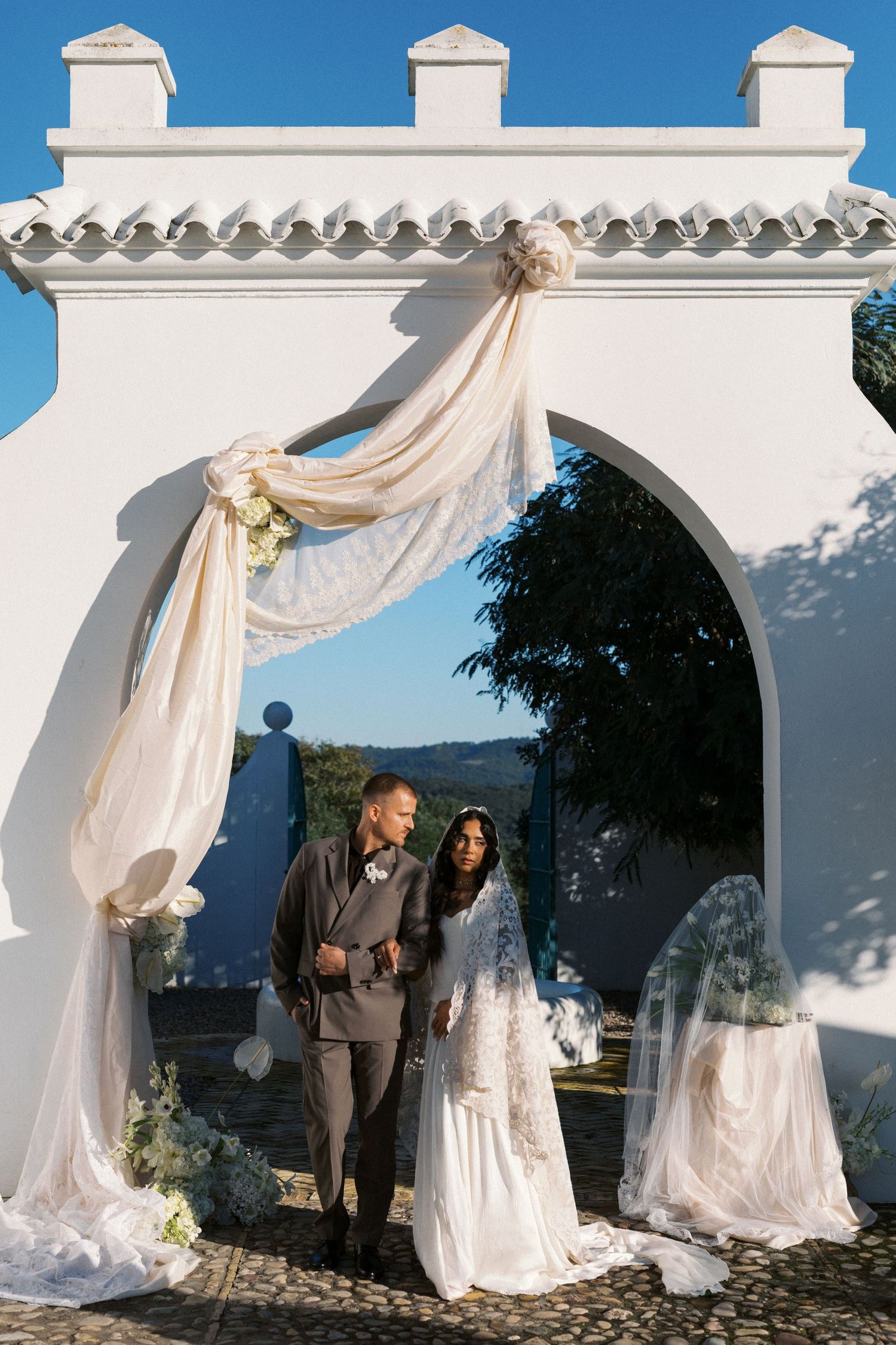 Bride and groom walk through white arched Mediterranean gate decorated with draped fabric and floral arrangements.