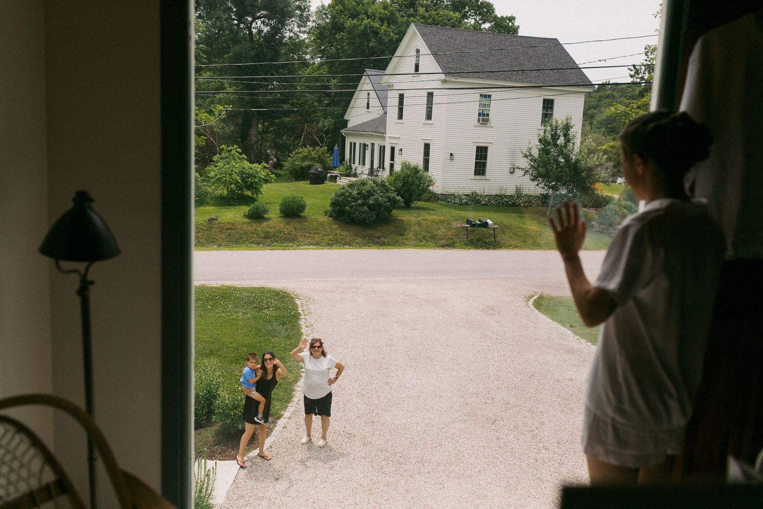Person waves from doorway at two people standing in driveway of white farmhouse on sunny day.