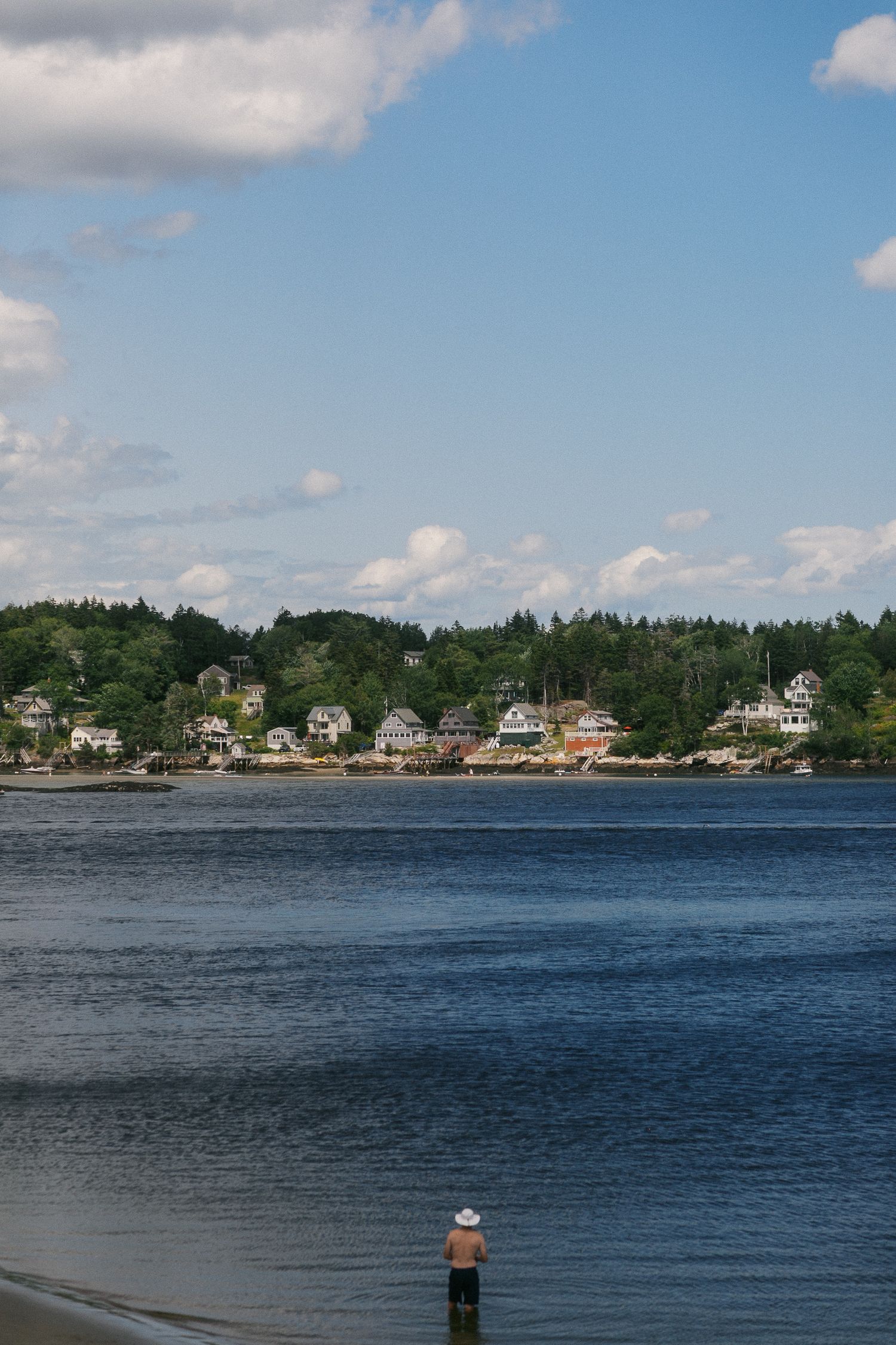 Person wading in calm blue water with a charming coastal Maine village and lush green trees under a partly cloudy sky.