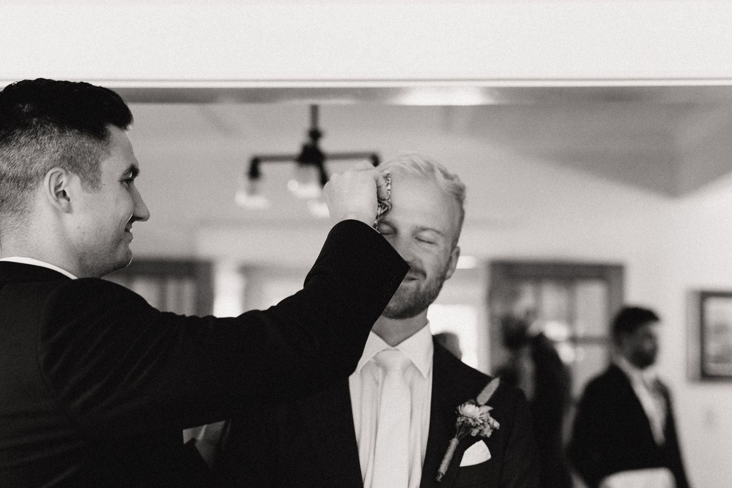 A black and white candid moment shows two men in formal attire sharing a joyful interaction at a wedding.