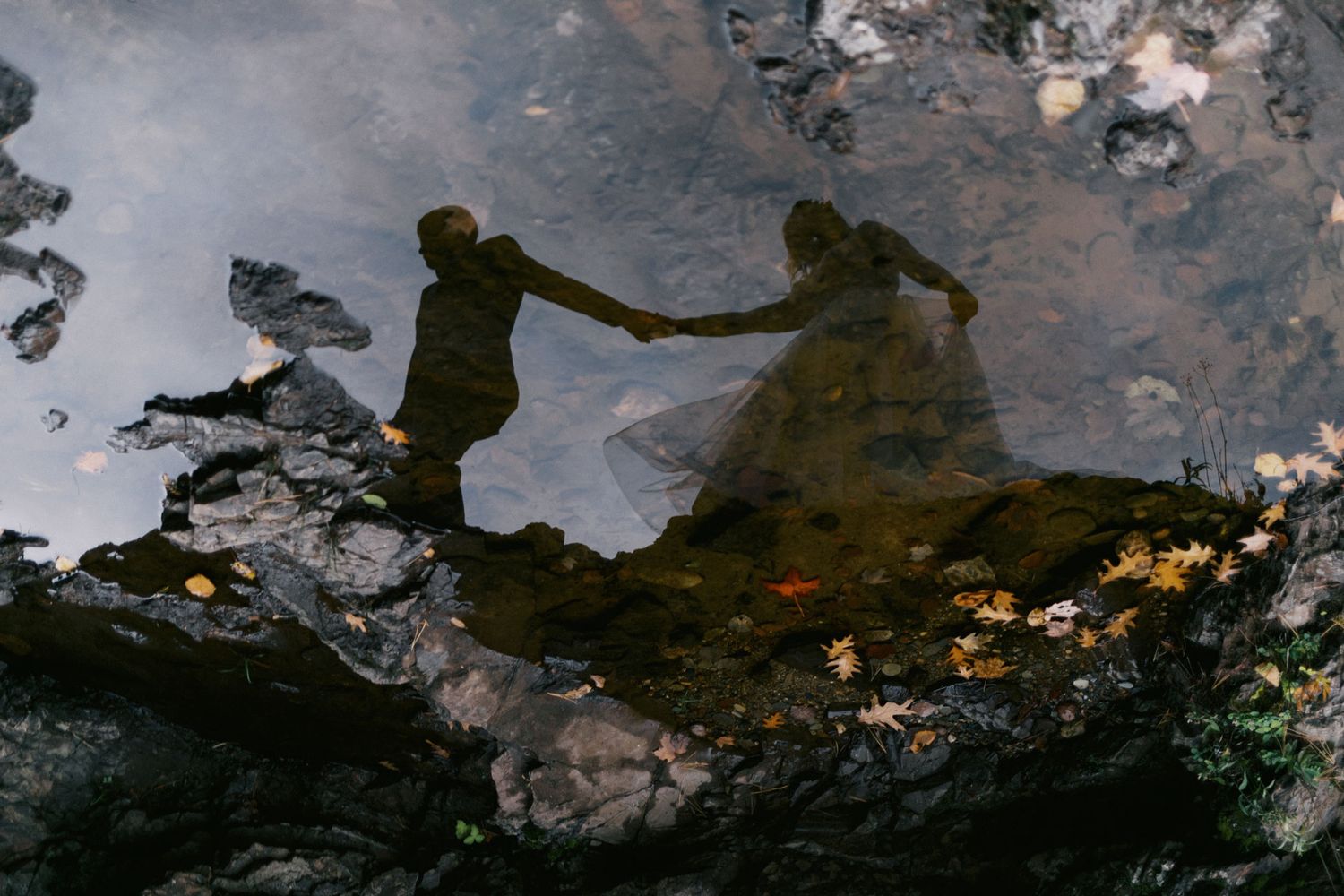 Romantic reflection of couple holding hands in shallow water with autumn leaves and rocks.