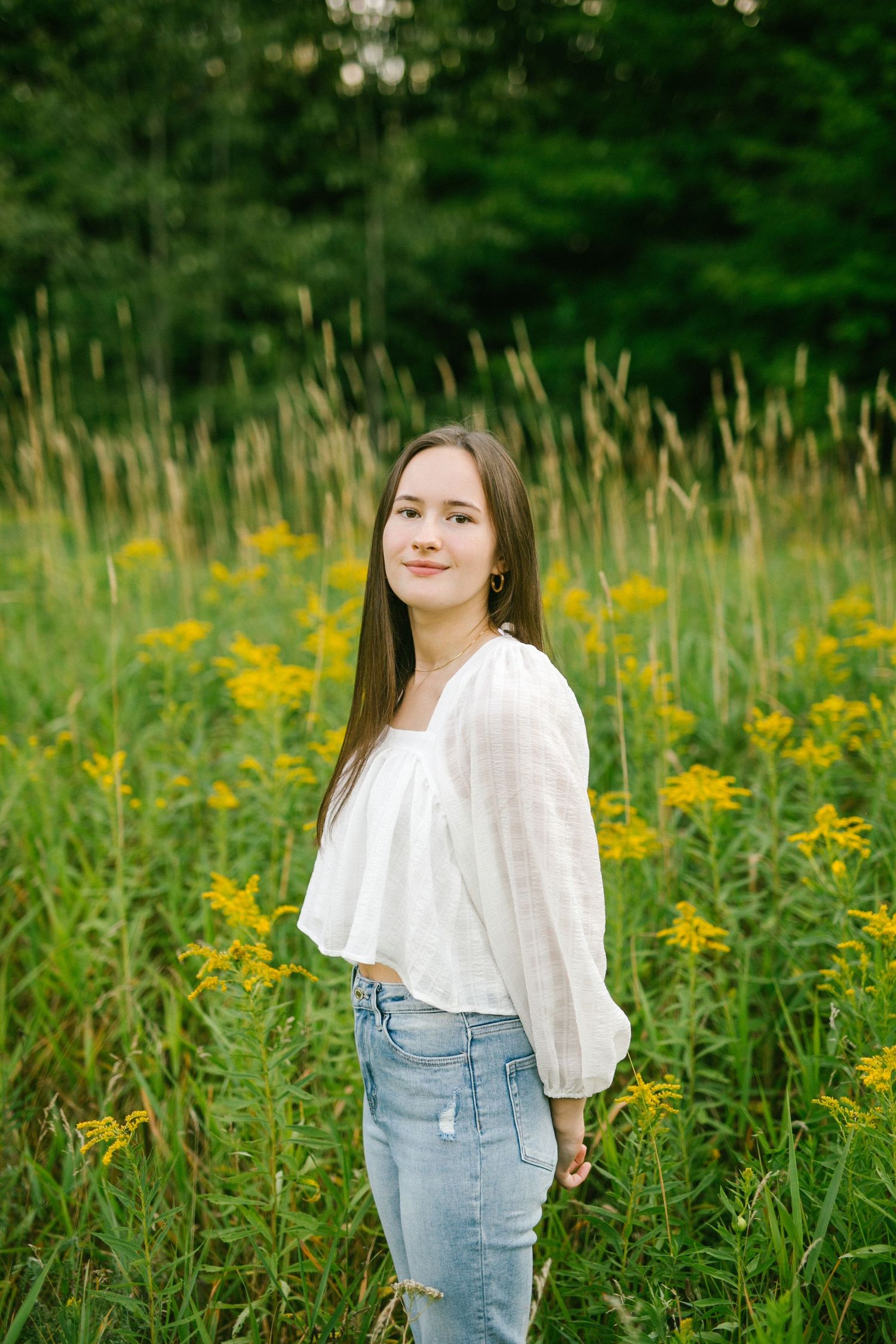 Young brunette woman in white blouse and jeans stands smiling among tall yellow wildflowers in a green field.