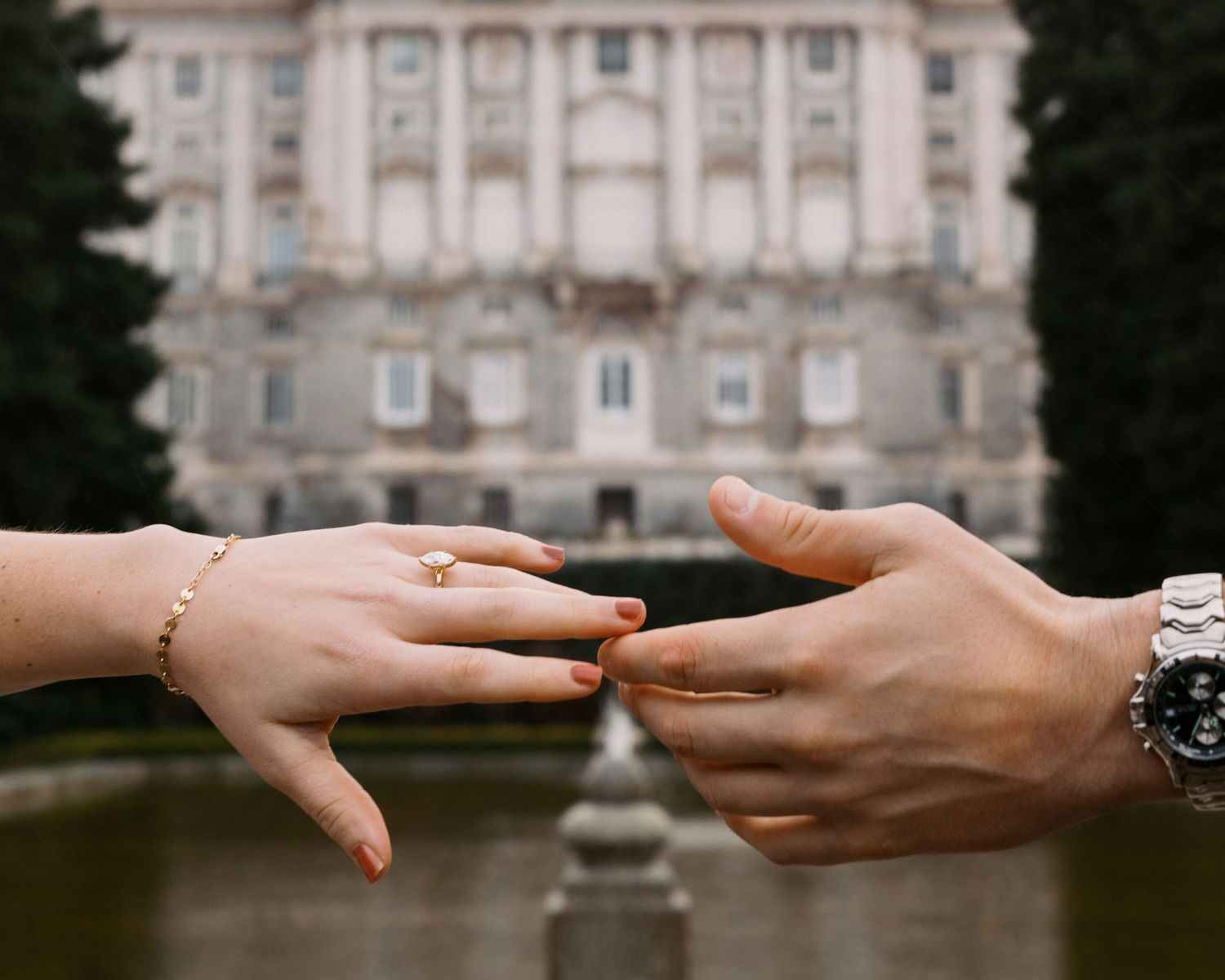 Close-up of couple's hands touching with engagement ring near a grand historic building and fountain.