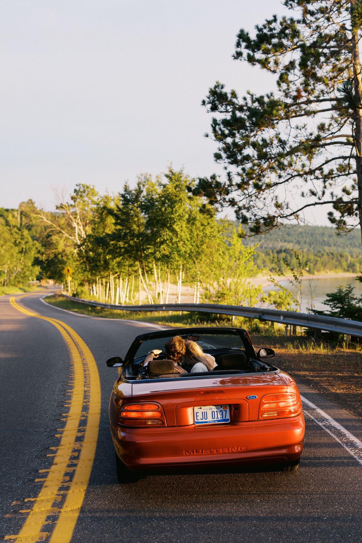 Red convertible car on a scenic winding road surrounded by lush green trees and a lake in golden hour light.