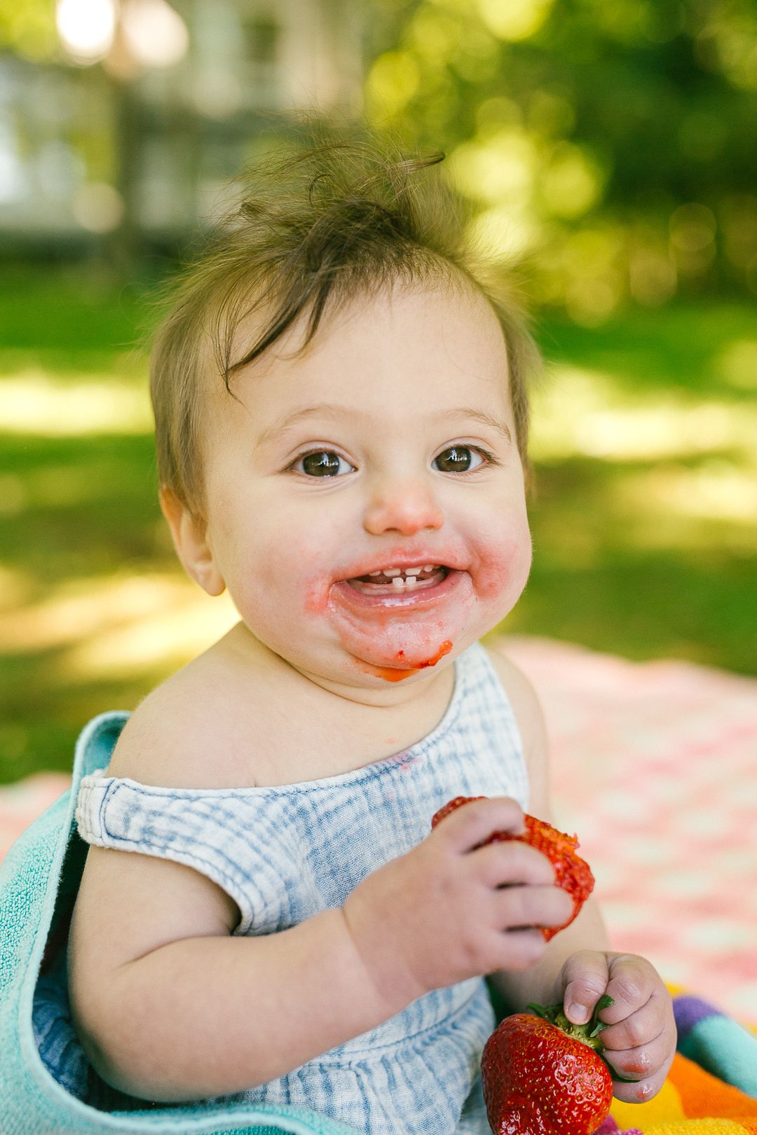 A happy baby girl with a messy strawberry-stained face holds a strawberry outdoors in the sun.
