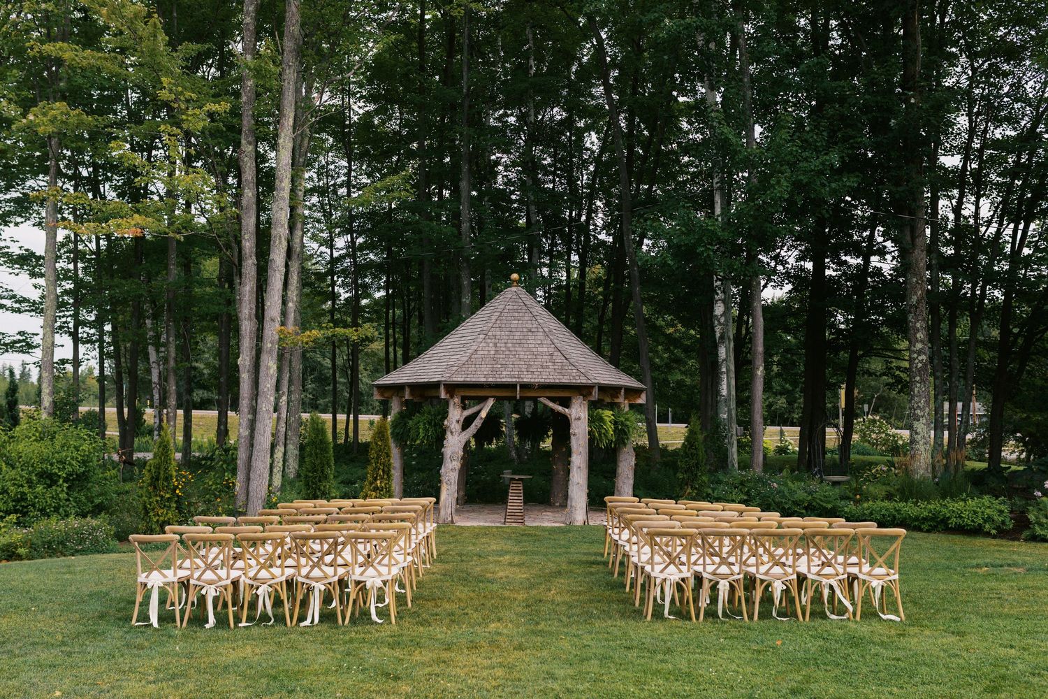 Outdoor wedding ceremony setup with wooden chairs and a rustic gazebo altar surrounded by tall trees on a green lawn.