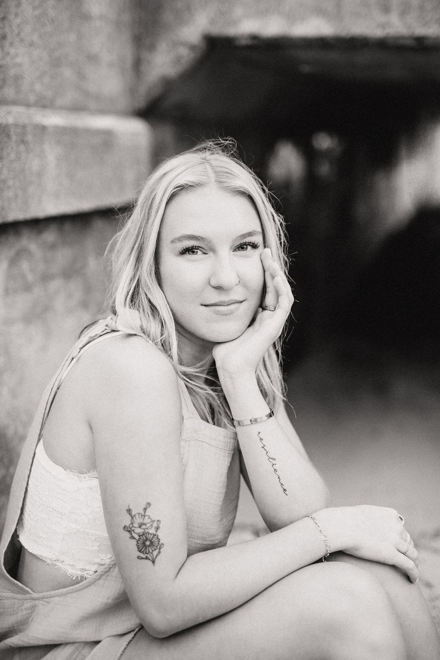 Black and white portrait of a blonde young woman with a floral arm tattoo sitting and smiling softly outdoors.