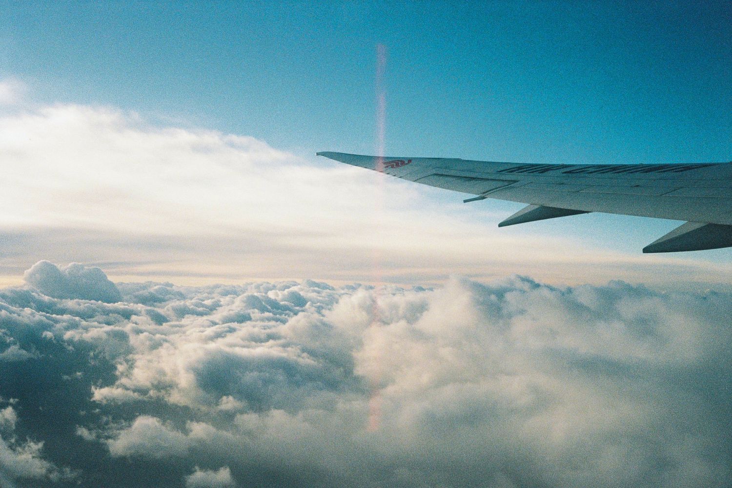 Aerial view of airplane wing above fluffy white clouds under a bright blue sky.