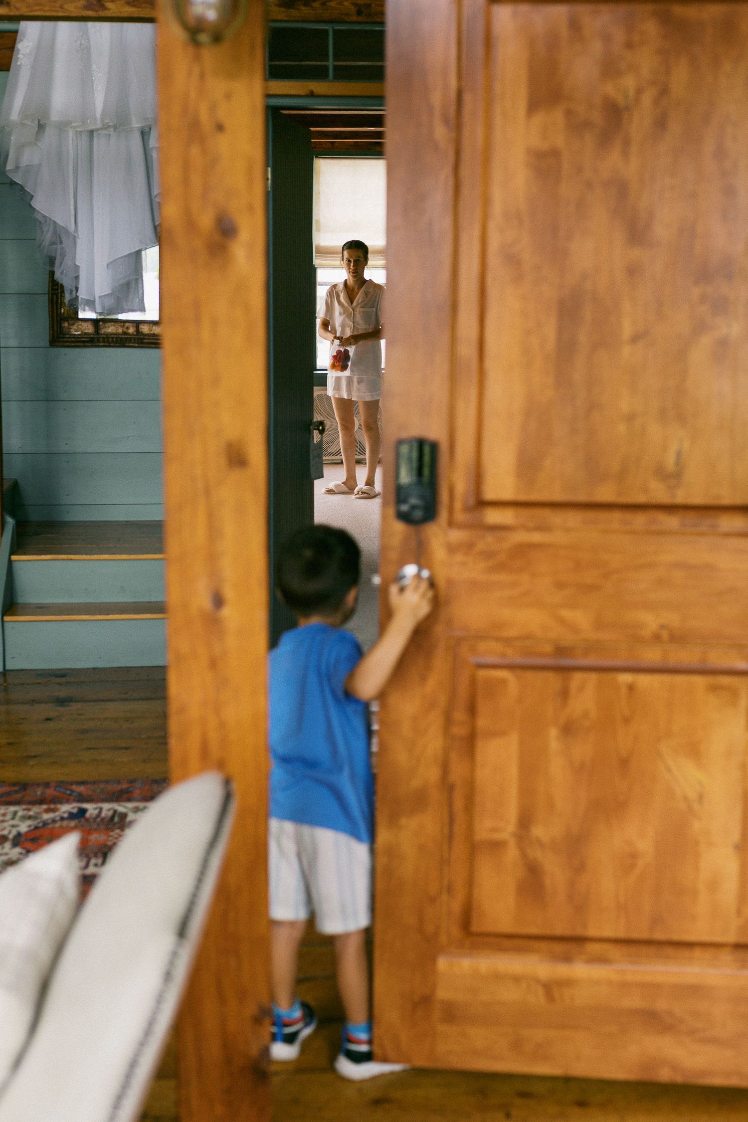 Young boy in blue shirt opening a wooden door while a woman stands in the background of a rustic home.